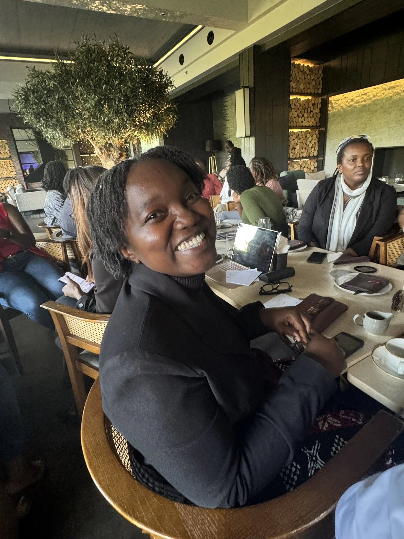 A woman smiling at a round table in a restaurant, with a group of people in the background. The setting is warmly lit with a tree and artistic wood paneling on the wall.