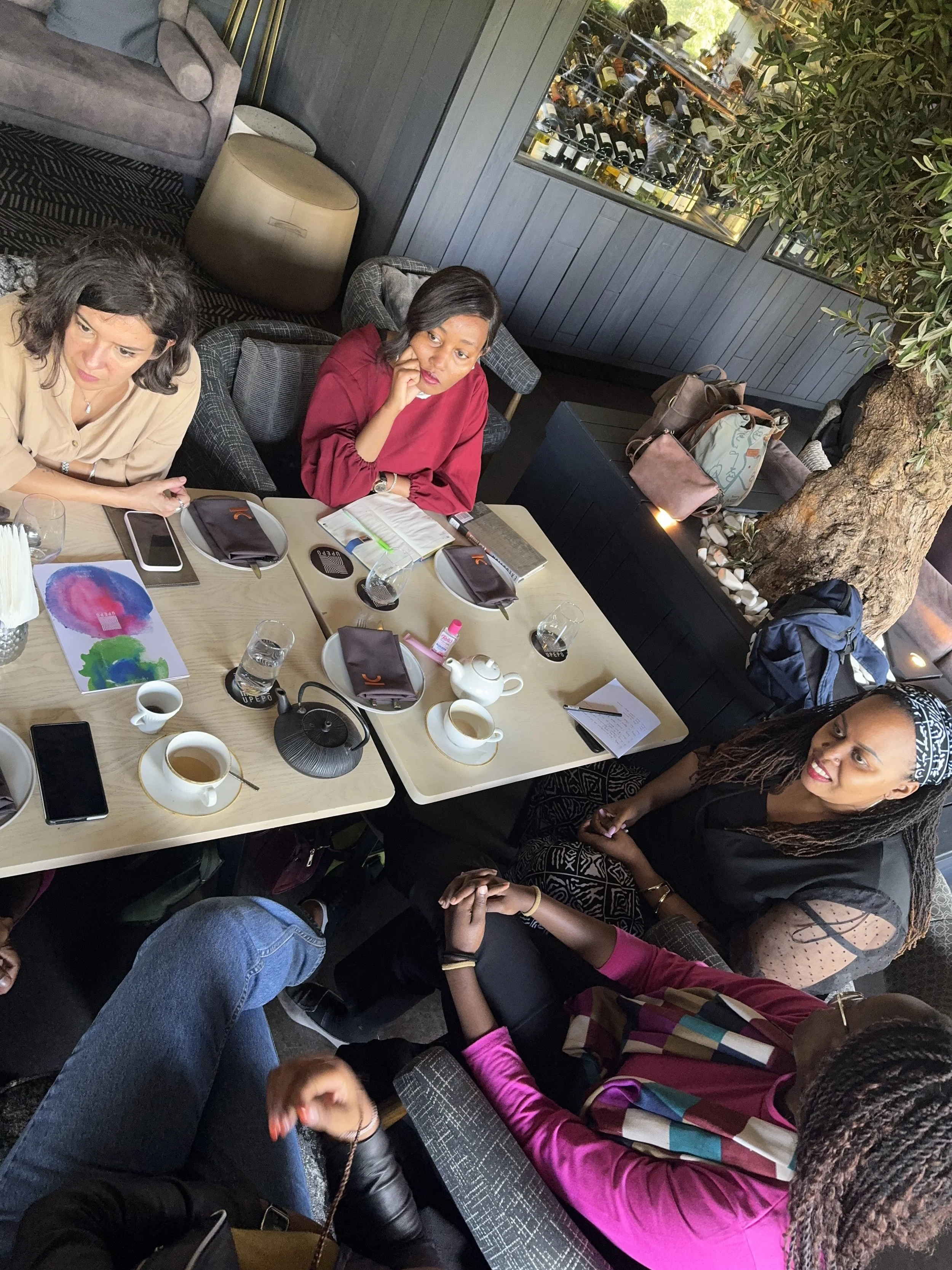 People sitting around a table in a restaurant, engaging in conversation. The table has drinks, notebooks, and miscellaneous items. A wine display is visible in the background, with a large tree integrated into the decor.