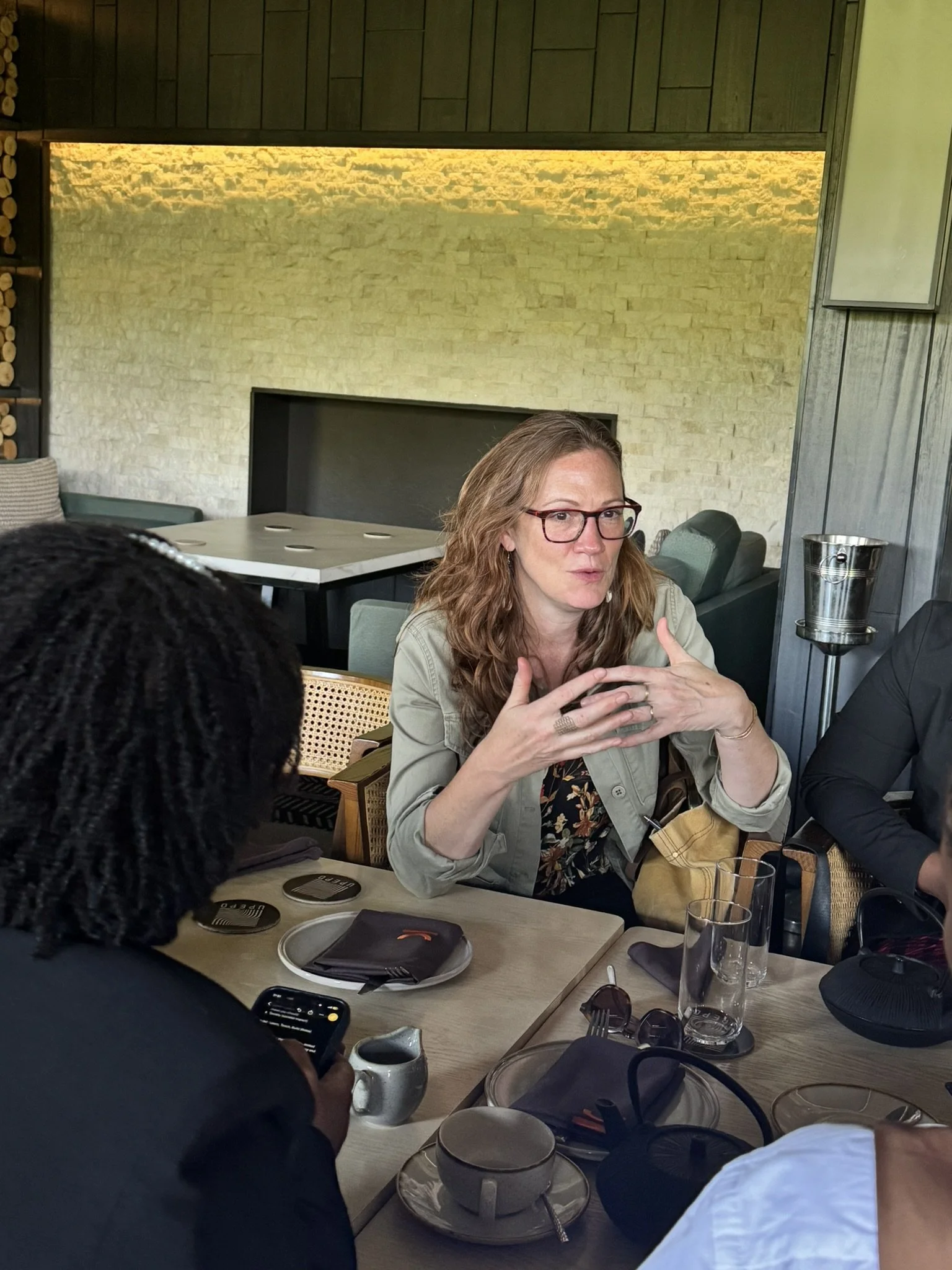 A woman with glasses and long brown hair sitting at a dining table in a restaurant, engaged in conversation. The table has plates, glasses, napkins, and utensils. The background shows a textured tan brick wall and wooden paneling.