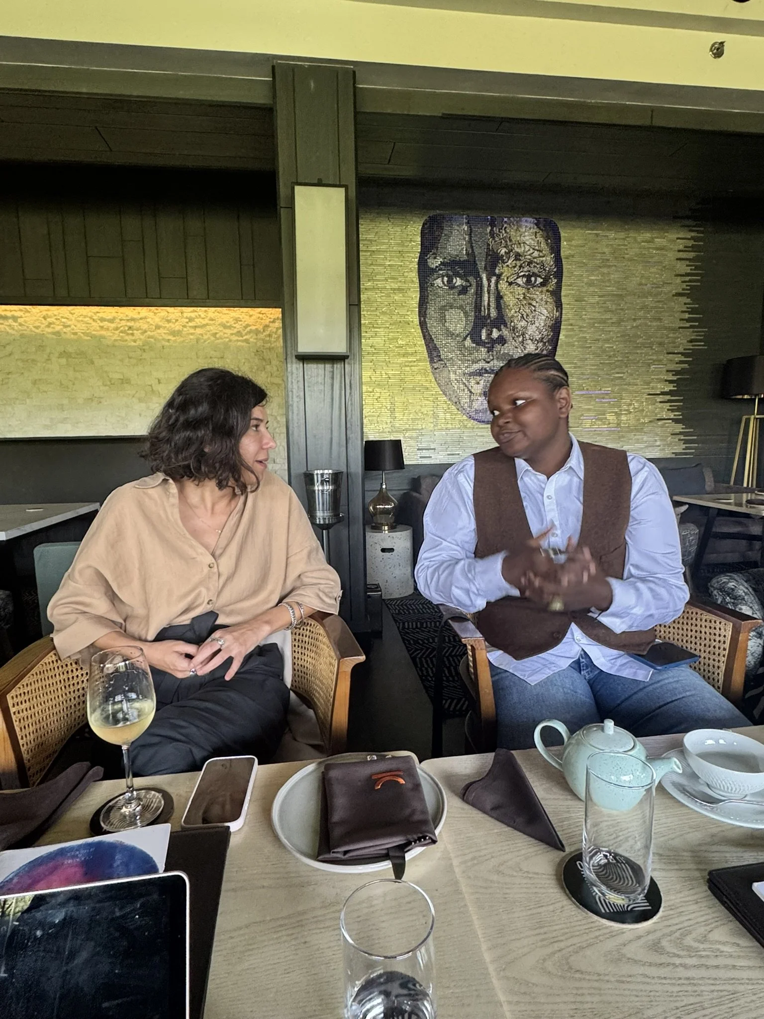 Two people sitting at a restaurant table engaging in conversation. The table has a wine glass, a teapot, and a smartphone. In the background, there is a large artistic wall piece resembling a face. The setting appears stylish and cozy.