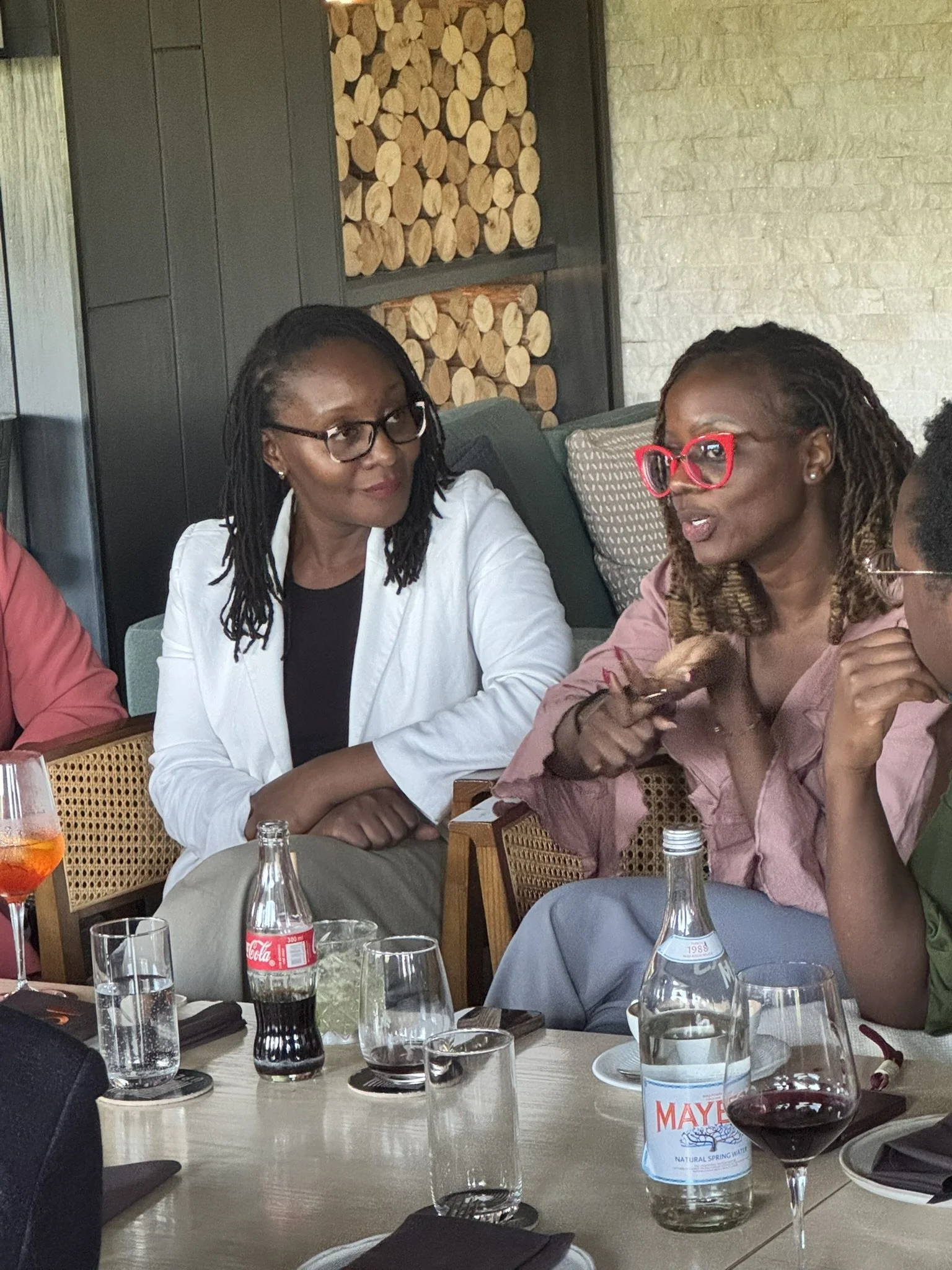 Two women sitting at a table in a restaurant, engaged in conversation. One wears glasses and a white blazer, the other wears large red glasses and a pink shirt. The table has various drinks, including a bottle of Meywasser spring water, wine, and sod