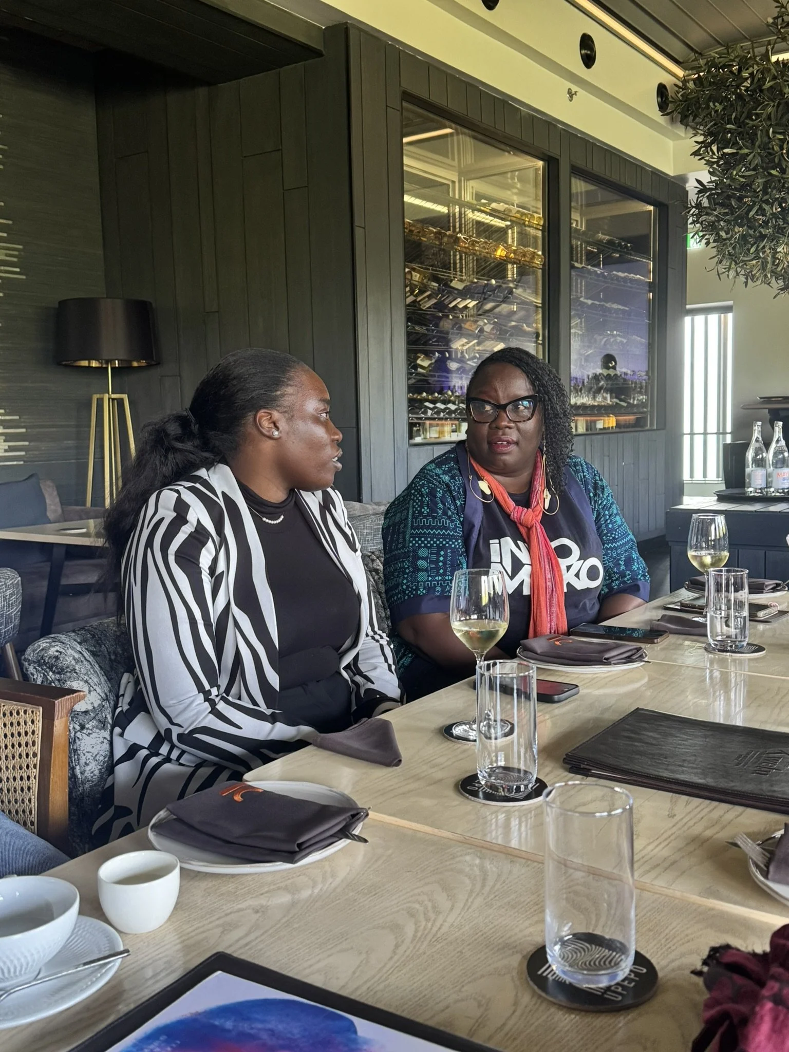 Two women sitting at a restaurant table having a conversation. The woman on the left has dark hair tied back and is wearing a black and white striped blazer. The woman on the right has curly hair, glasses, and is wearing a colorful jacket and a red s