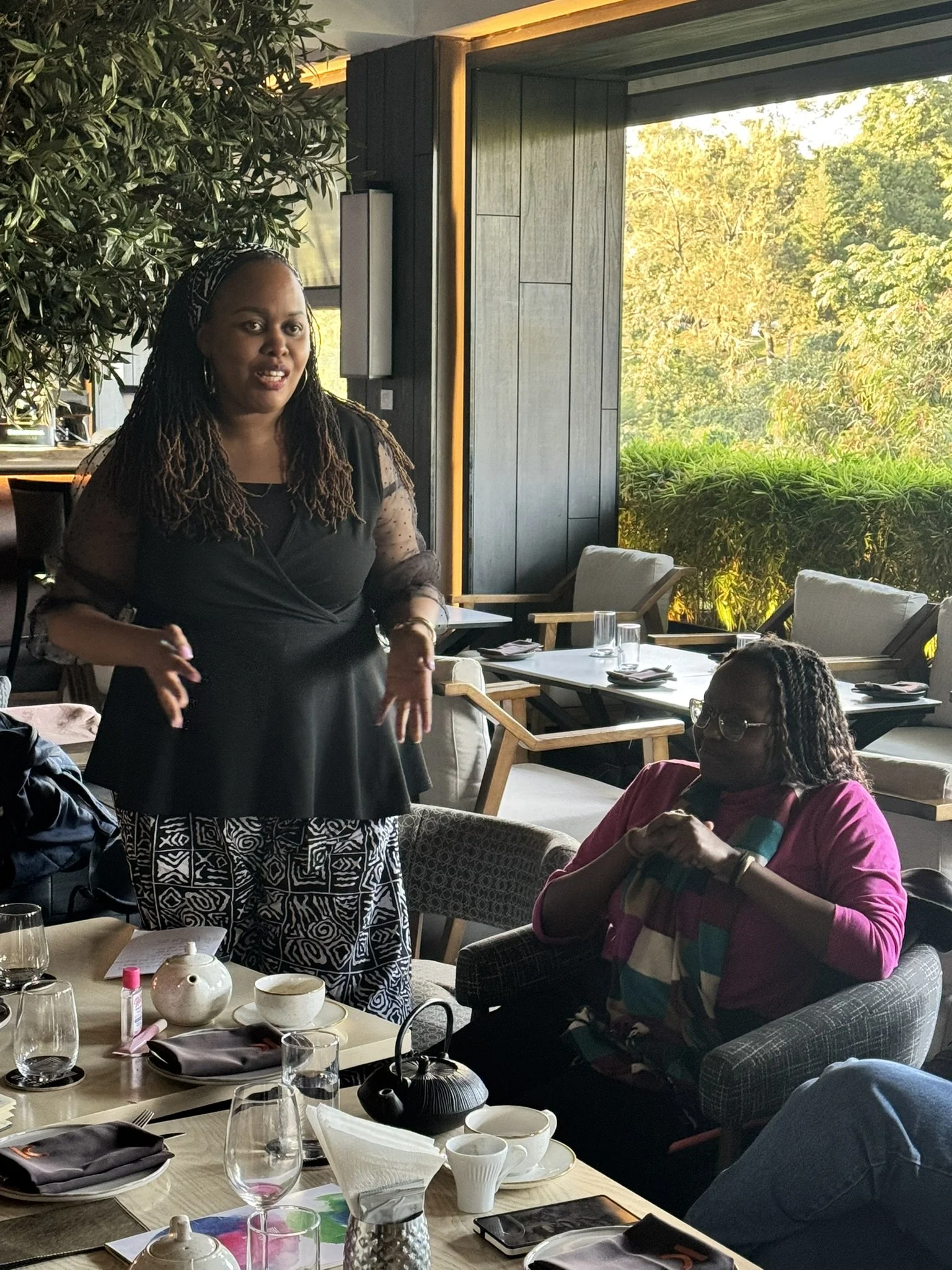 A woman with dreadlocks wearing a black top and patterned pants standing and talking in a restaurant with large windows showing trees outside. Seated woman wearing glasses and a pink shirt is listening. The table has plates, glasses, a teapot, and va