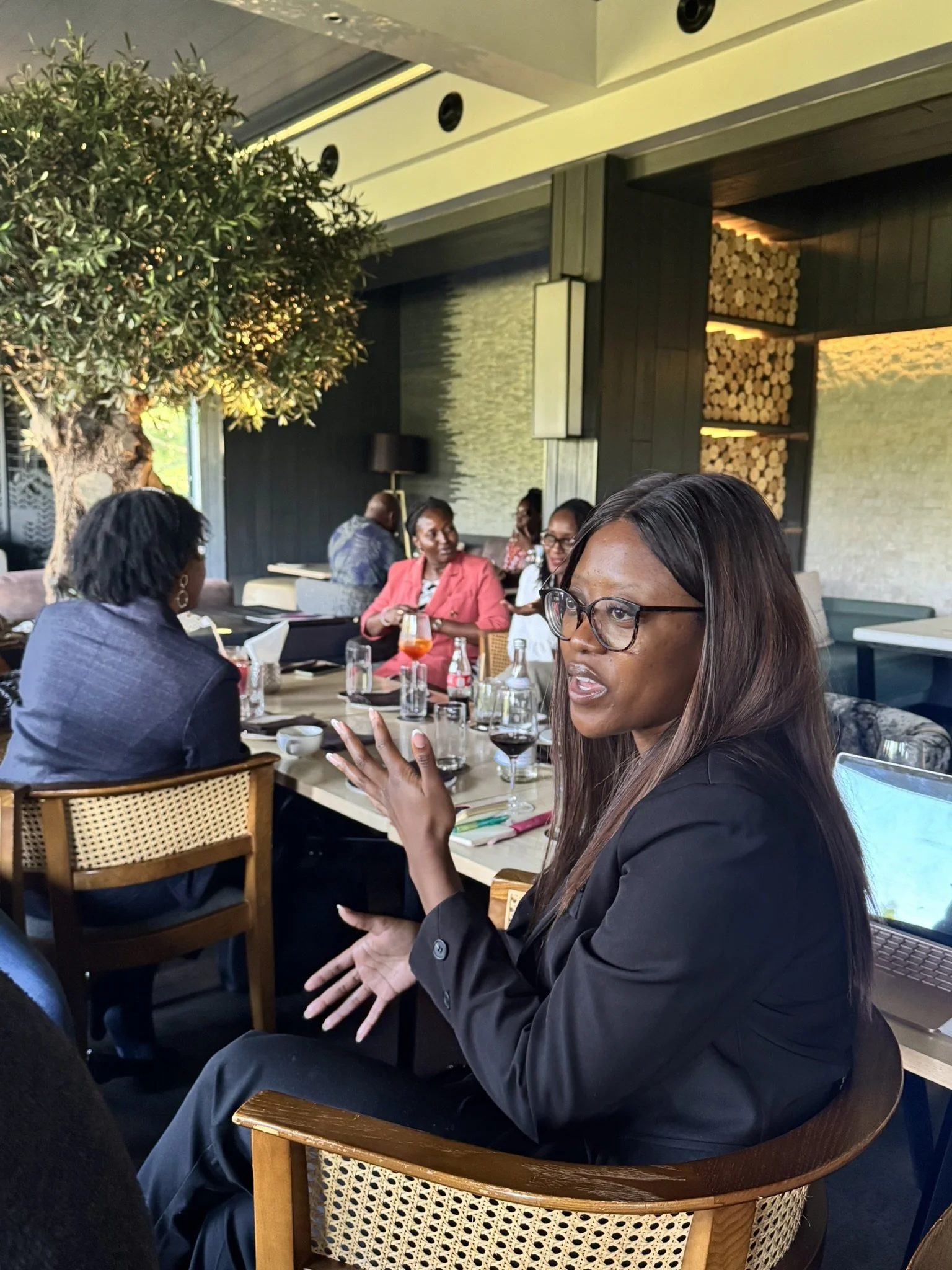 People sitting at a restaurant table, engaging in conversation. A woman in the foreground is speaking, wearing glasses and a black blazer. The restaurant has modern decor with a large tree, wood accents, and warm lighting.