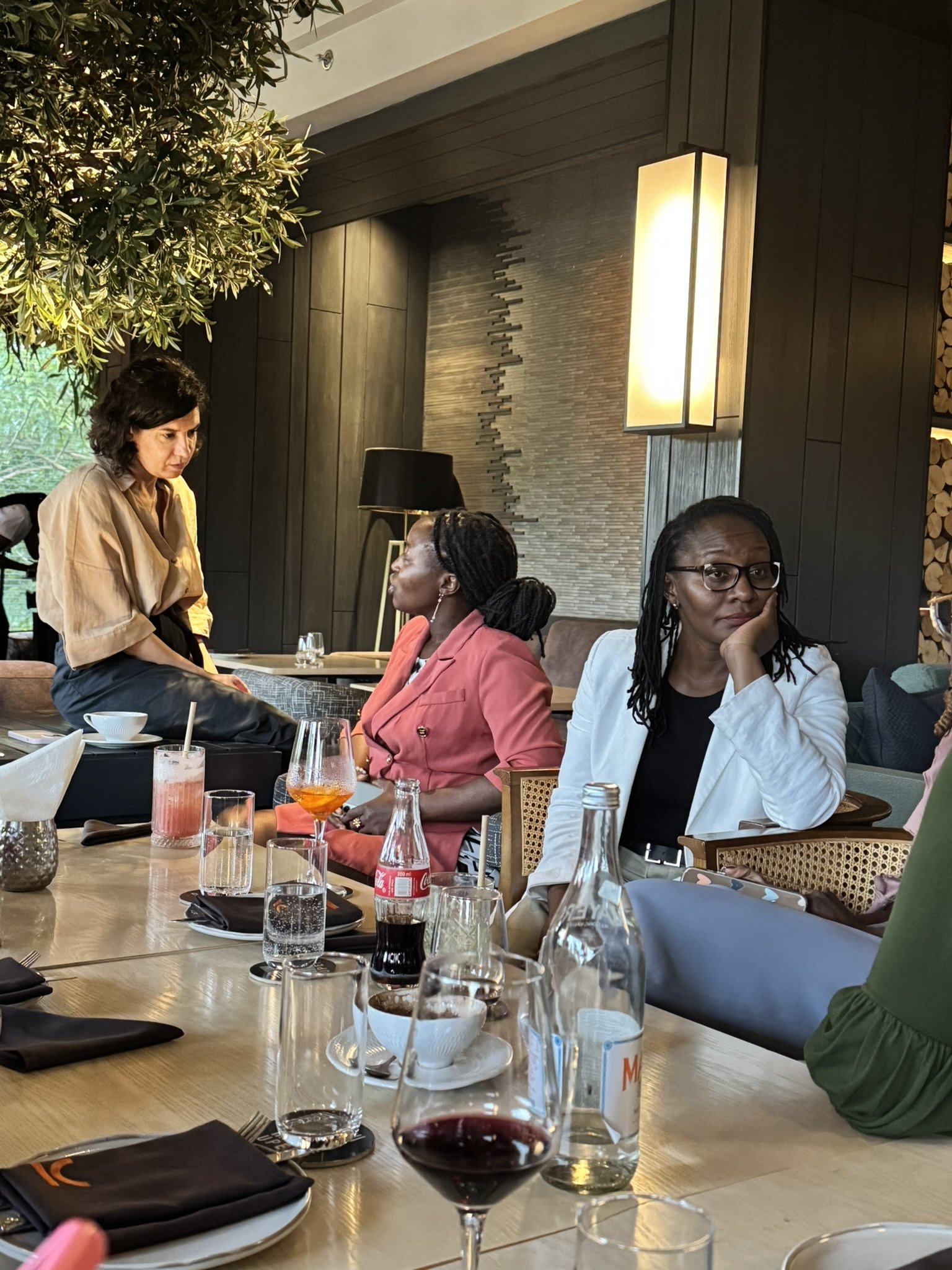 A group of people seated around a dining table with drinks and tableware, engaging in conversation inside a restaurant with dark wood paneling, a textured brick wall, and warm lighting.