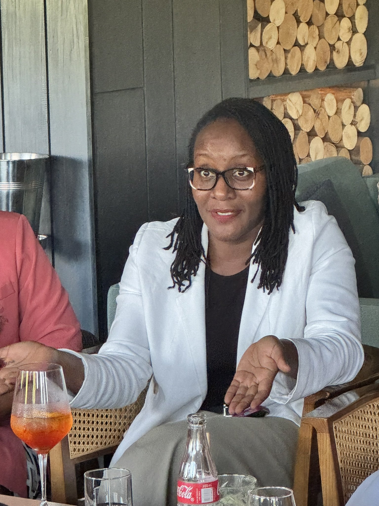 A woman with glasses and long, braided hair, wearing a white jacket and black shirt, sitting at a table in a restaurant. There are glasses and a Coca-Cola bottle on the table. The background shows a wall with stacked firewood.