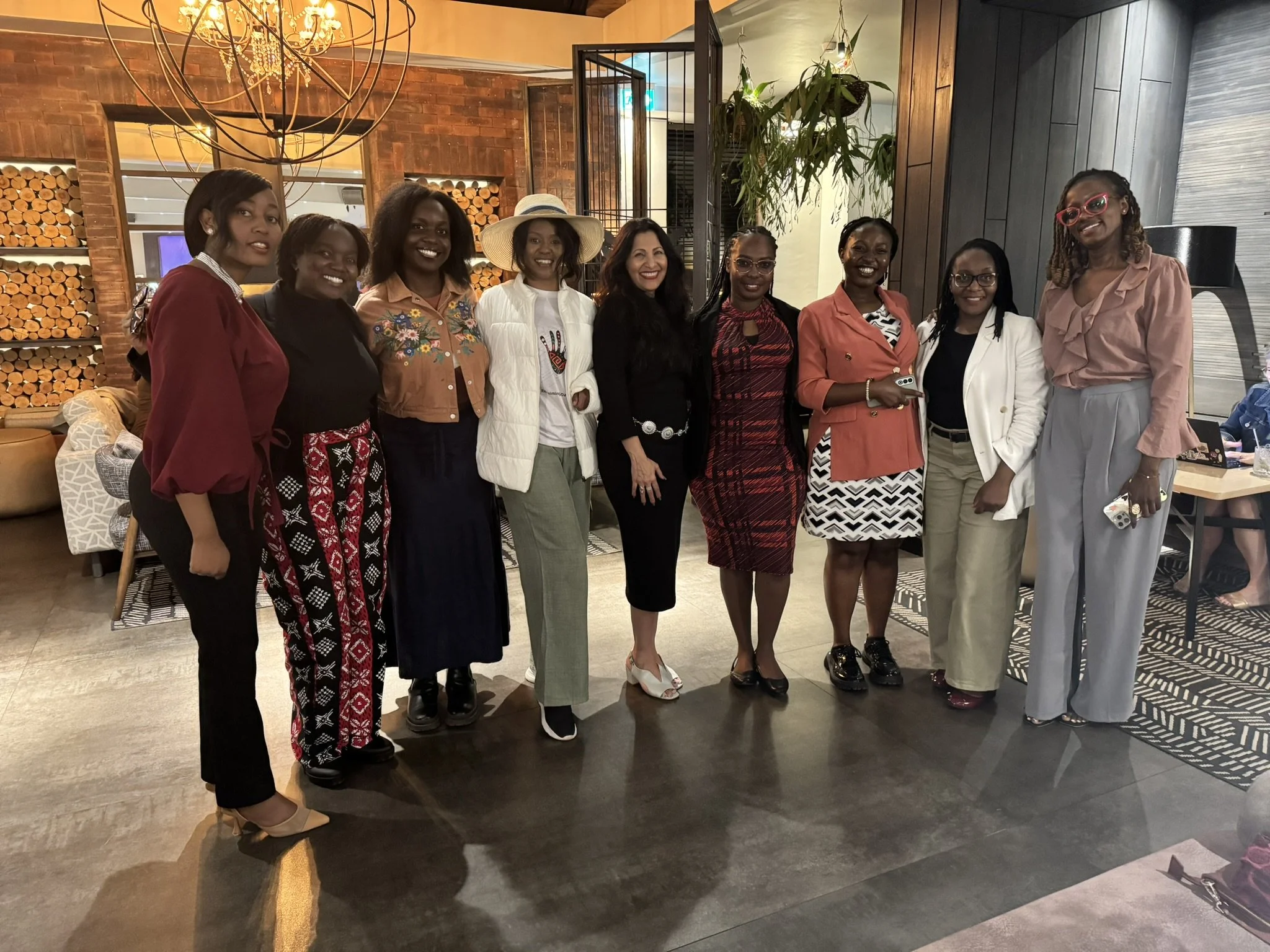 A group of ten women standing together indoors, smiling for a photo. They are dressed in colorful and patterned outfits. The background features a brick wall, a chandelier, and some modern decor in a warmly lit restaurant or event space.