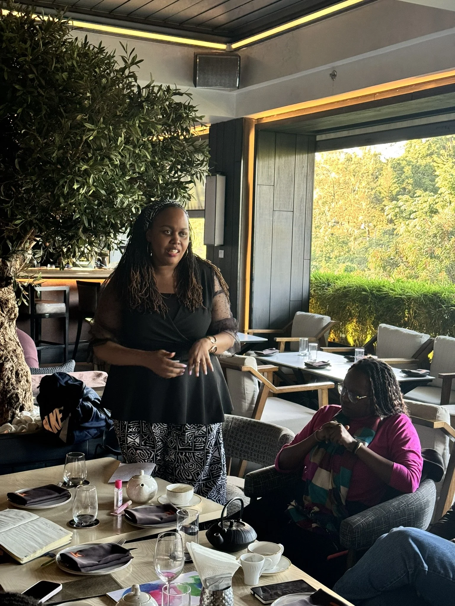 Women gathered in a restaurant, with one woman standing and speaking, while others are seated at a table with glasses, plates, and notebooks. Bright natural light from large windows illuminates the room, and greenery is visible outside.