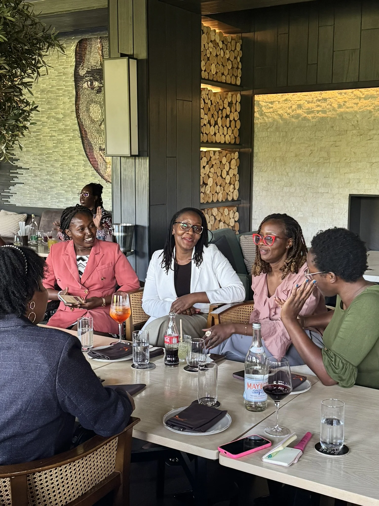 A group of women having a meeting or discussion in a restaurant or cafe. They are seated around a table with drinks, notebooks, and a phone. The background shows a modern interior with wood accents and decorative firewood storage.