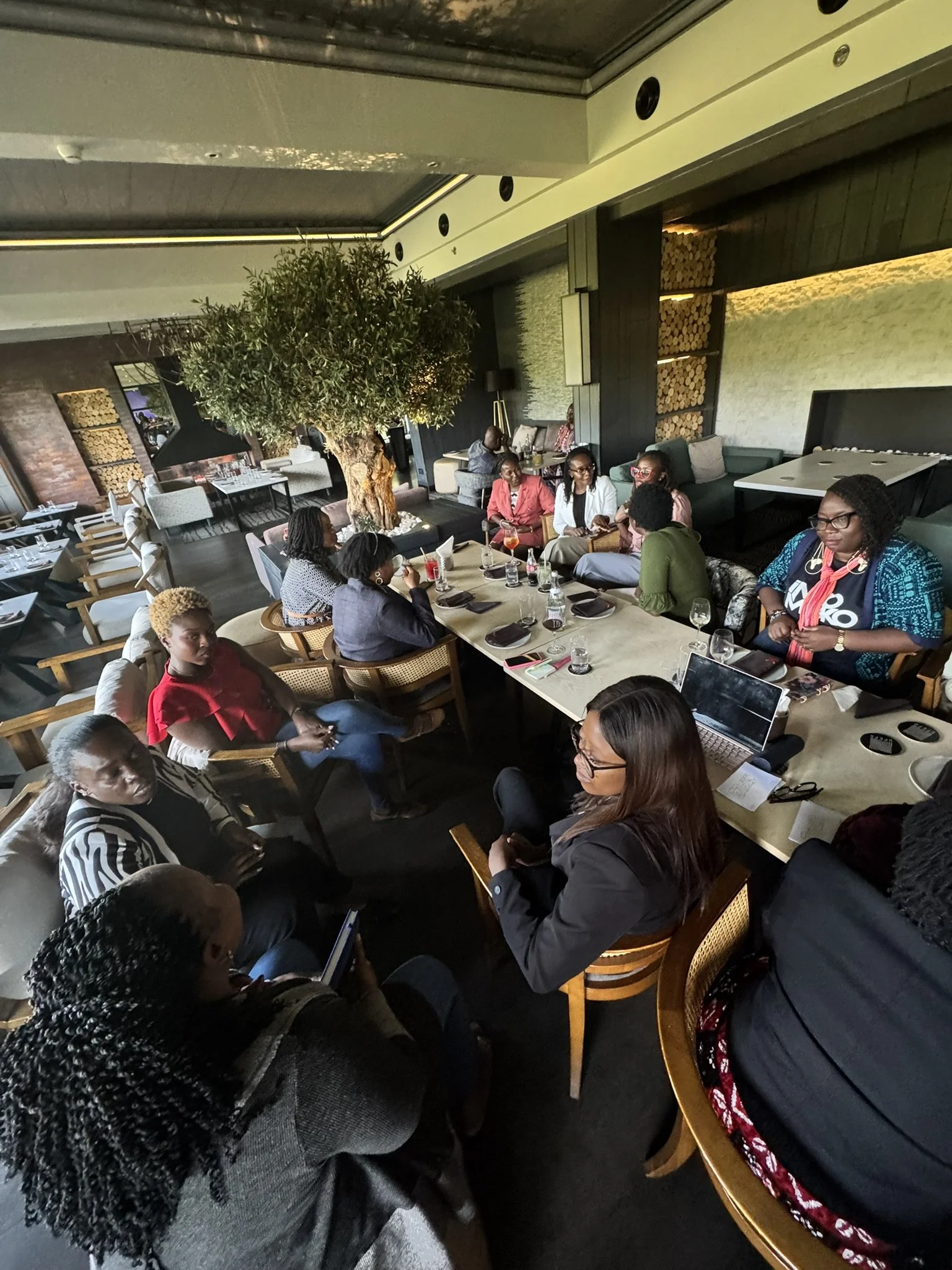 Group of women gathered around a large table in a modern restaurant or lounge with a tree in the center, engaging in conversation.