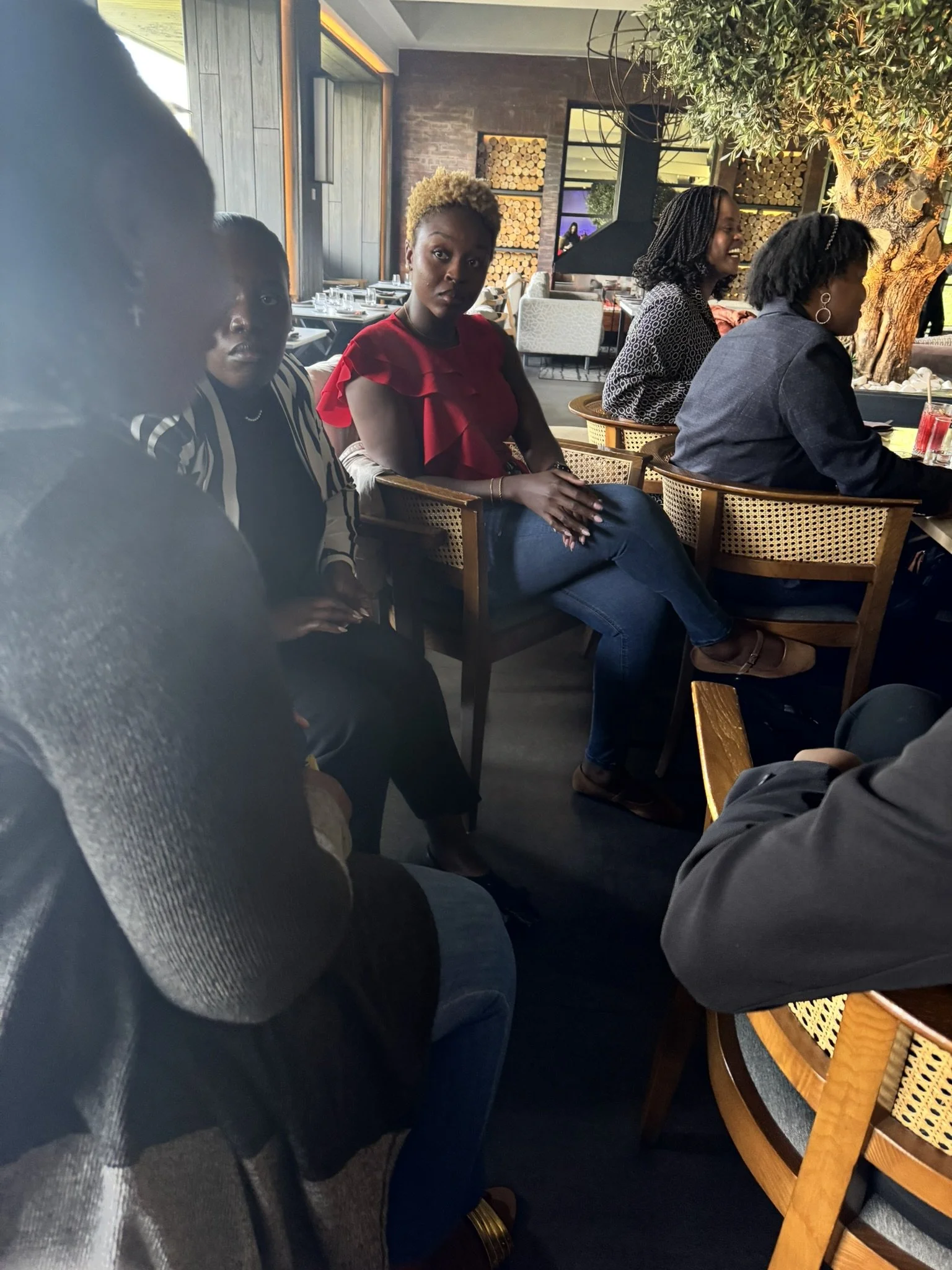 Four women sitting at a table in a restaurant, engaged in conversation, with a decorative tree and wood log art on the wall in the background.