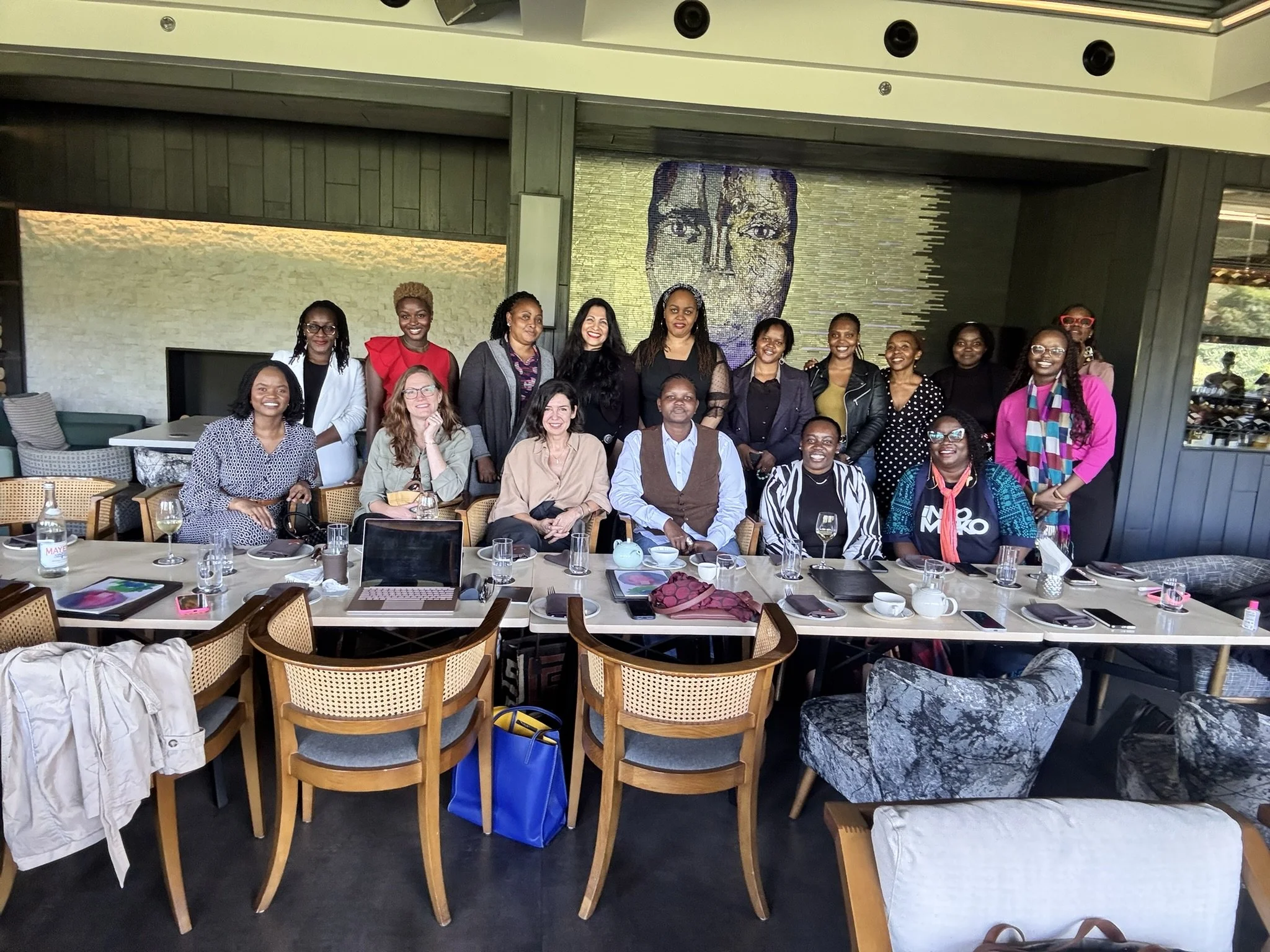 A group of women and one man gathered around a dining table in a restaurant or conference room, posing for a photo. The table is set with glasses, plates, and some items. The background features a large mosaic art piece of a face on the wall.