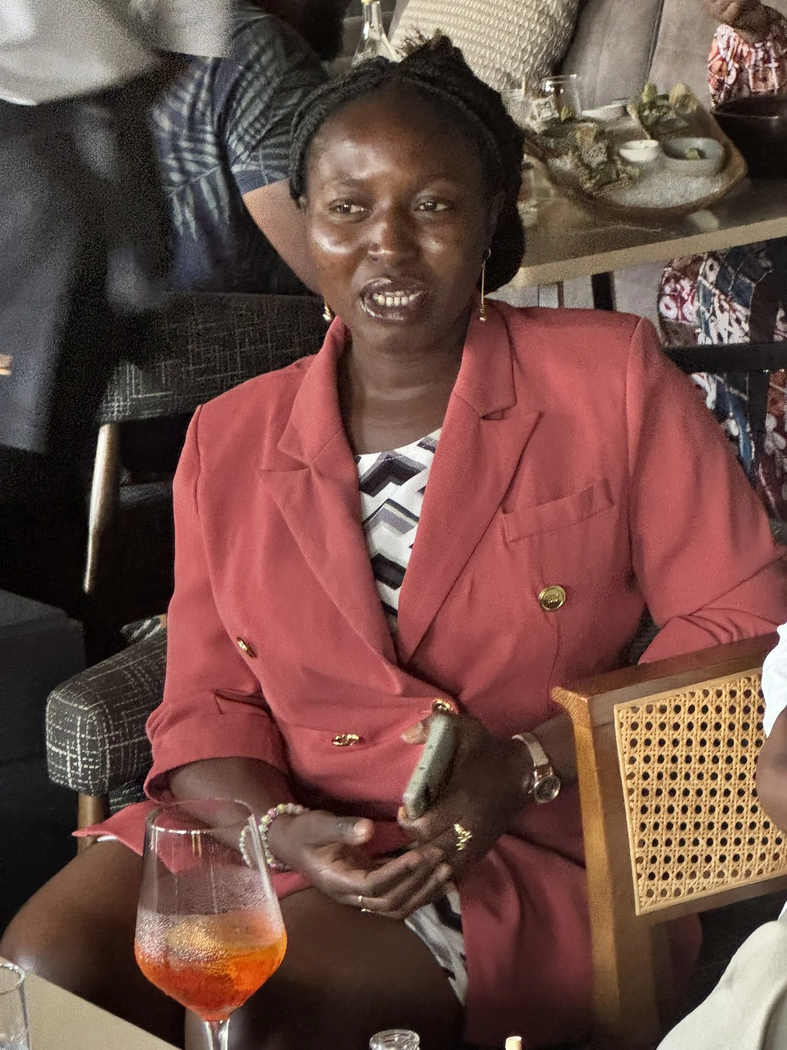 A woman wearing a red blazer and jewelry is sitting at a table in a restaurant with a glass of orange-colored beverage in front of her. She has dark hair styled in twists and is wearing earrings.