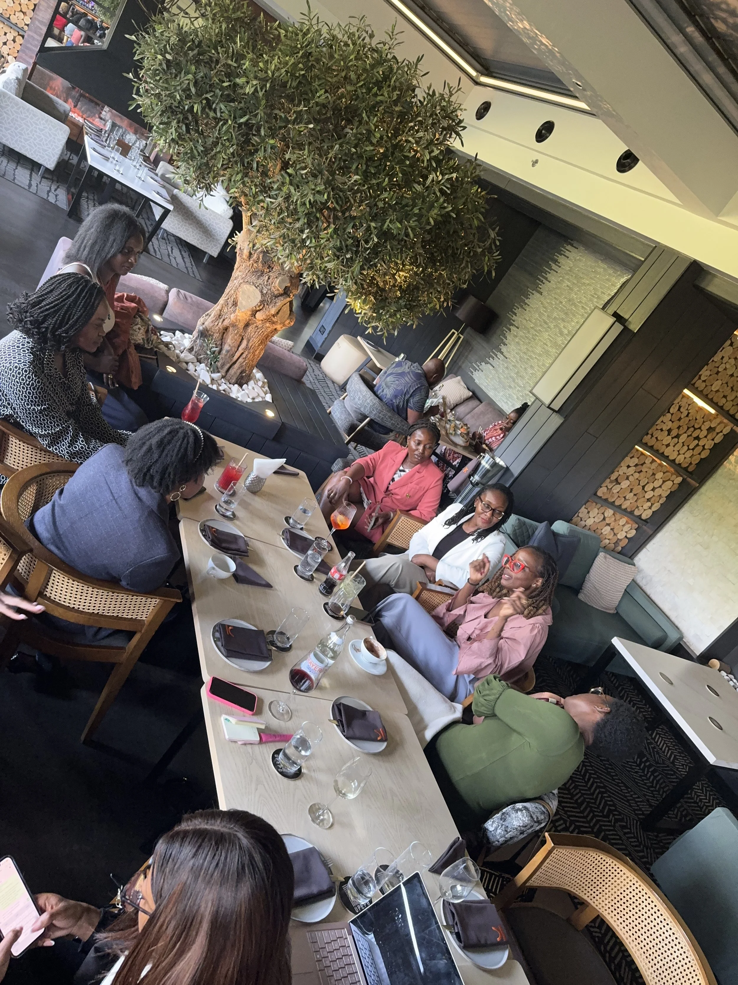 People sitting around a table in a restaurant with a large indoor tree, some using laptops and phones, drinks, and food on the table.