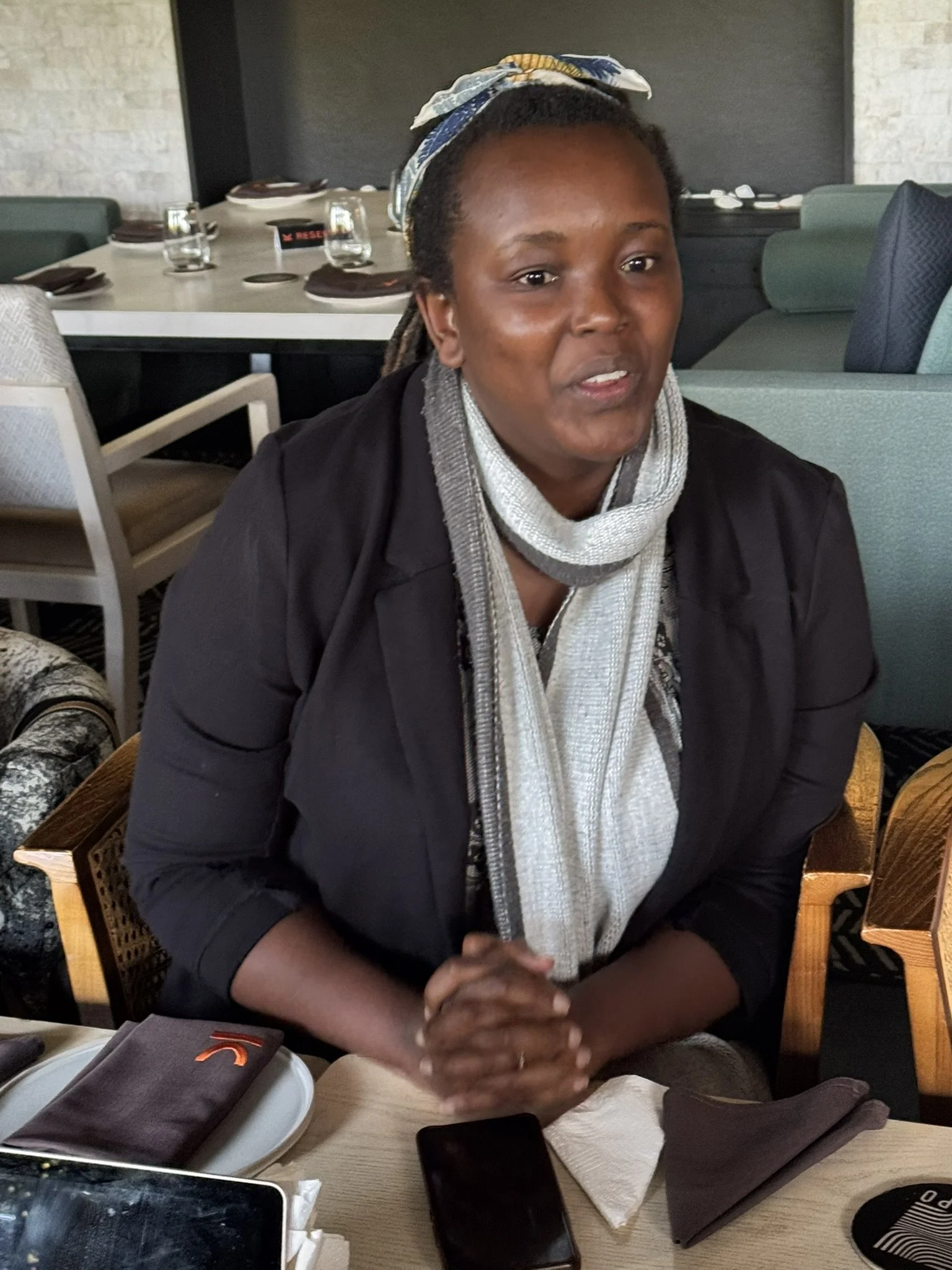 A woman with dark skin and braided hair, wearing a headband, seated at a restaurant table with a group of people, with plates, glasses, and personal belongings on the table.