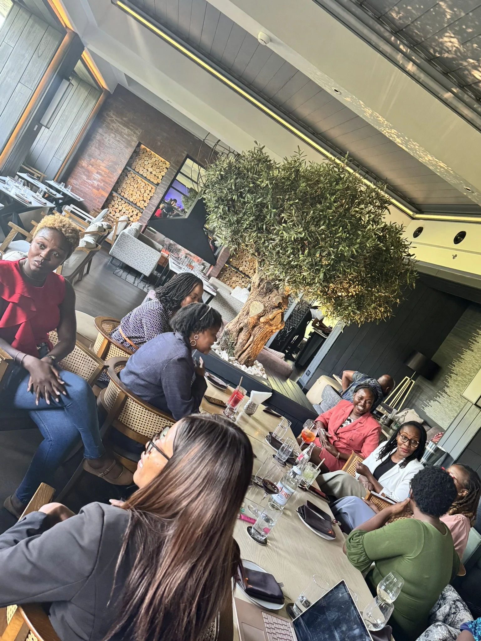 A group of people gathered around a dining table in a restaurant with a large indoor tree at the center, surrounded by wood and brick decor.