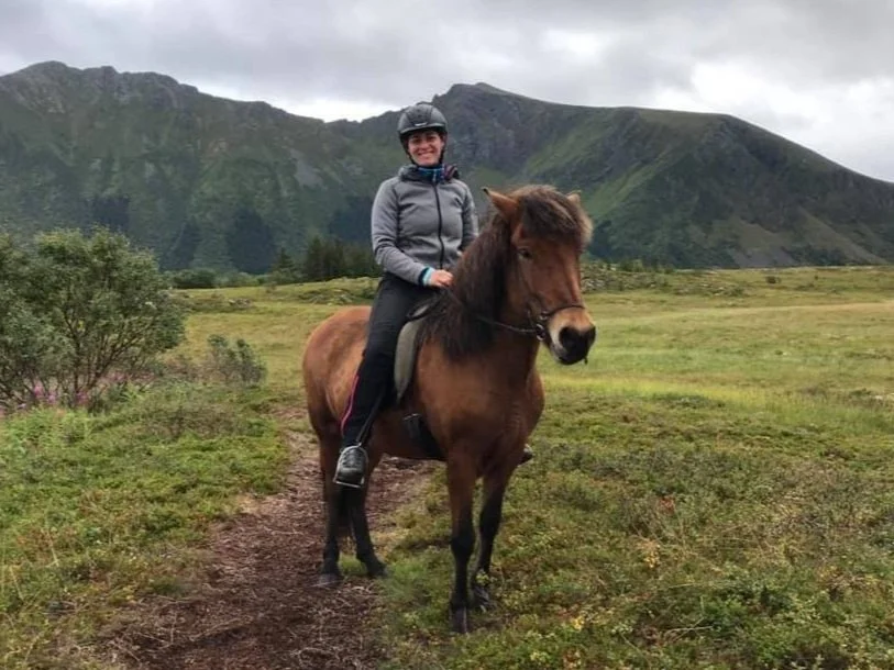 Eine Person reitet ein braunes Pferd auf einem Wanderweg in einer grünen, bergigen Landschaft. Die Person trägt einen Helm und lächelt. Der Himmel ist bewölkt.