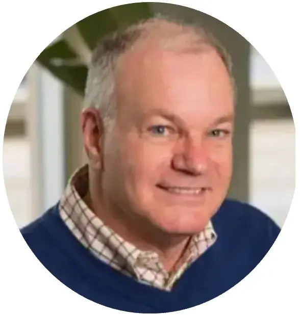 A middle-aged man with short gray hair smiling at the camera, wearing a checkered shirt and a navy blue sweater, with green plants in the background.