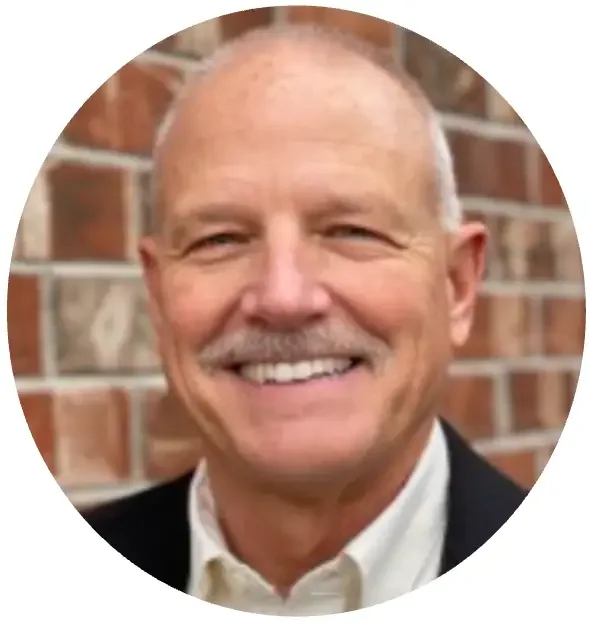 Close-up of a smiling middle-aged man with a mustache, wearing a white shirt and a dark jacket, standing in front of a brick wall.