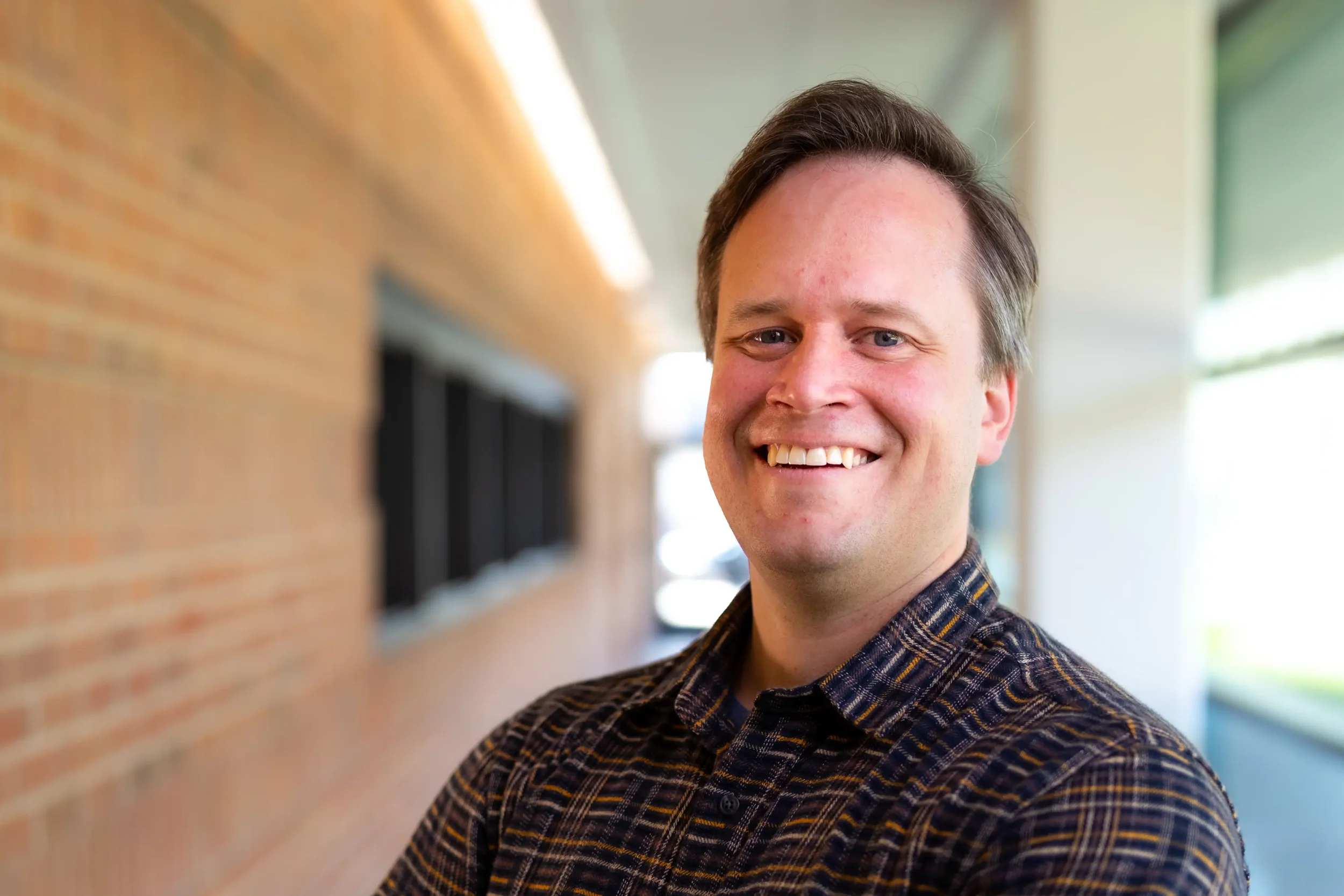 A smiling man with short brown hair and blue eyes, wearing a plaid shirt, standing outside near a brick building and windows.