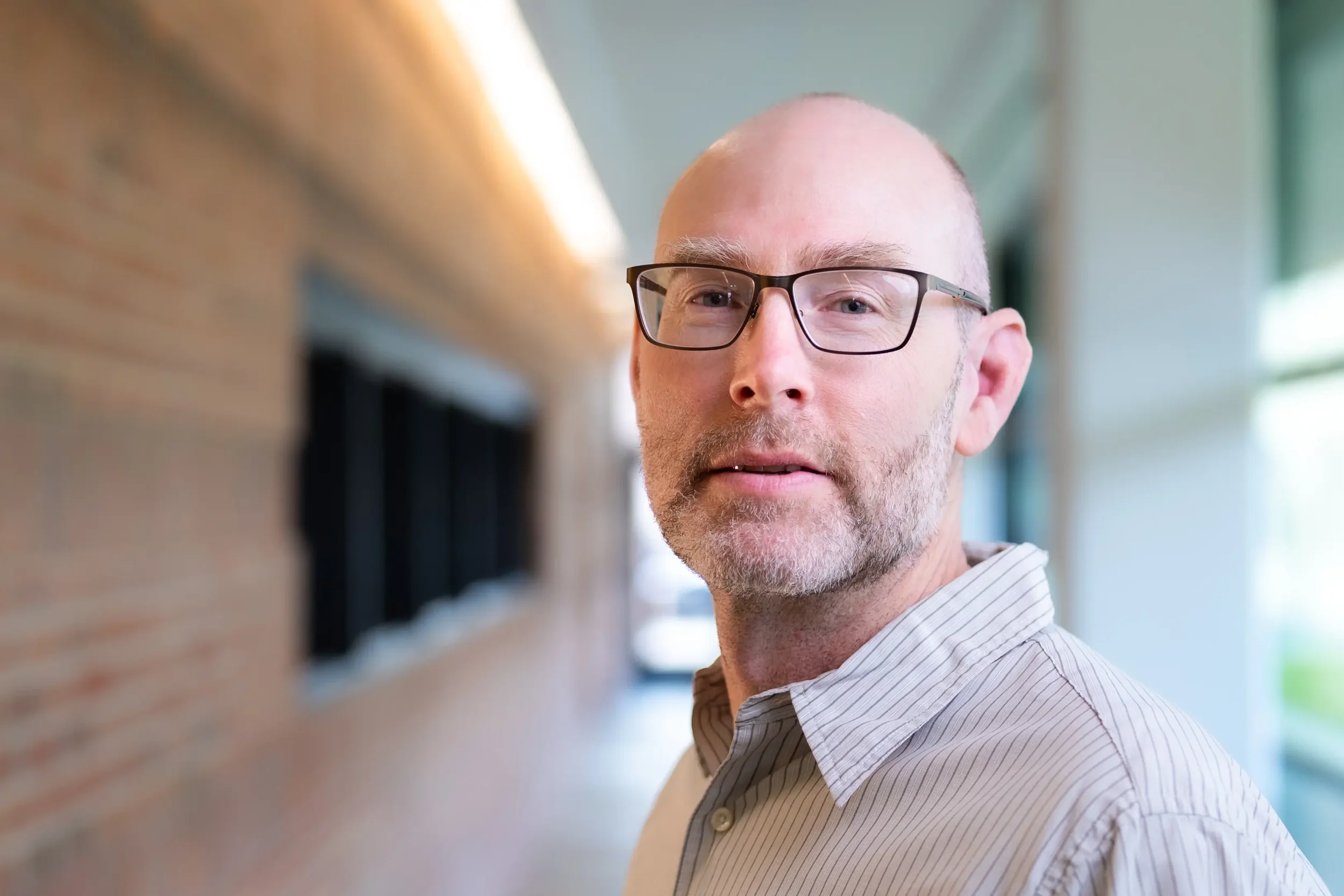 Close-up of a middle-aged man wearing glasses and a striped shirt, standing inside a building with large windows and a brick wall in the background.