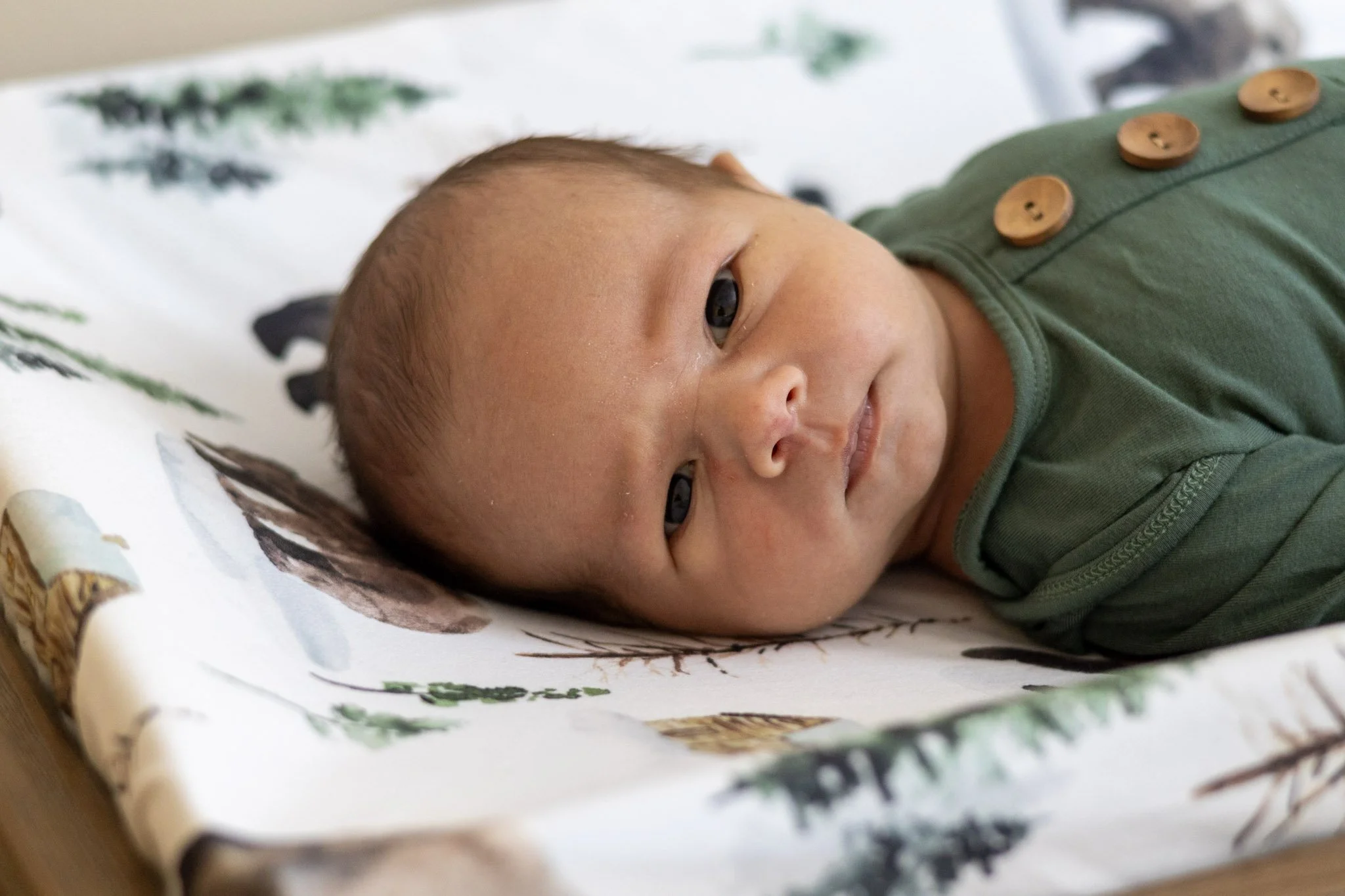 A baby lying on a white blanket with nature-themed illustrations, looking at the camera with a calm expression, wearing a green outfit with wooden buttons.