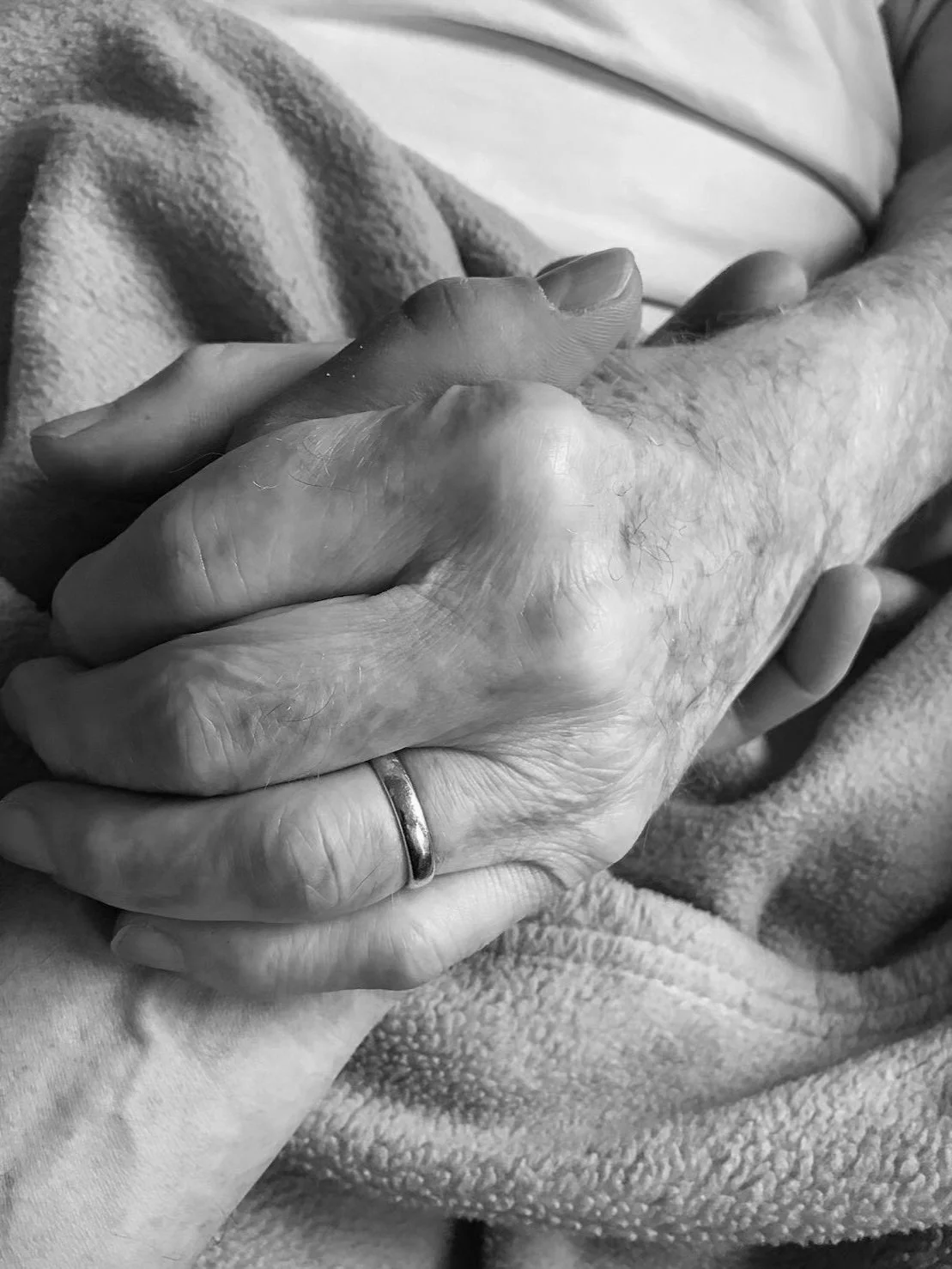 Close-up of two hands holding each other, one older and one younger, with one wearing a wedding band, on a soft blanket.