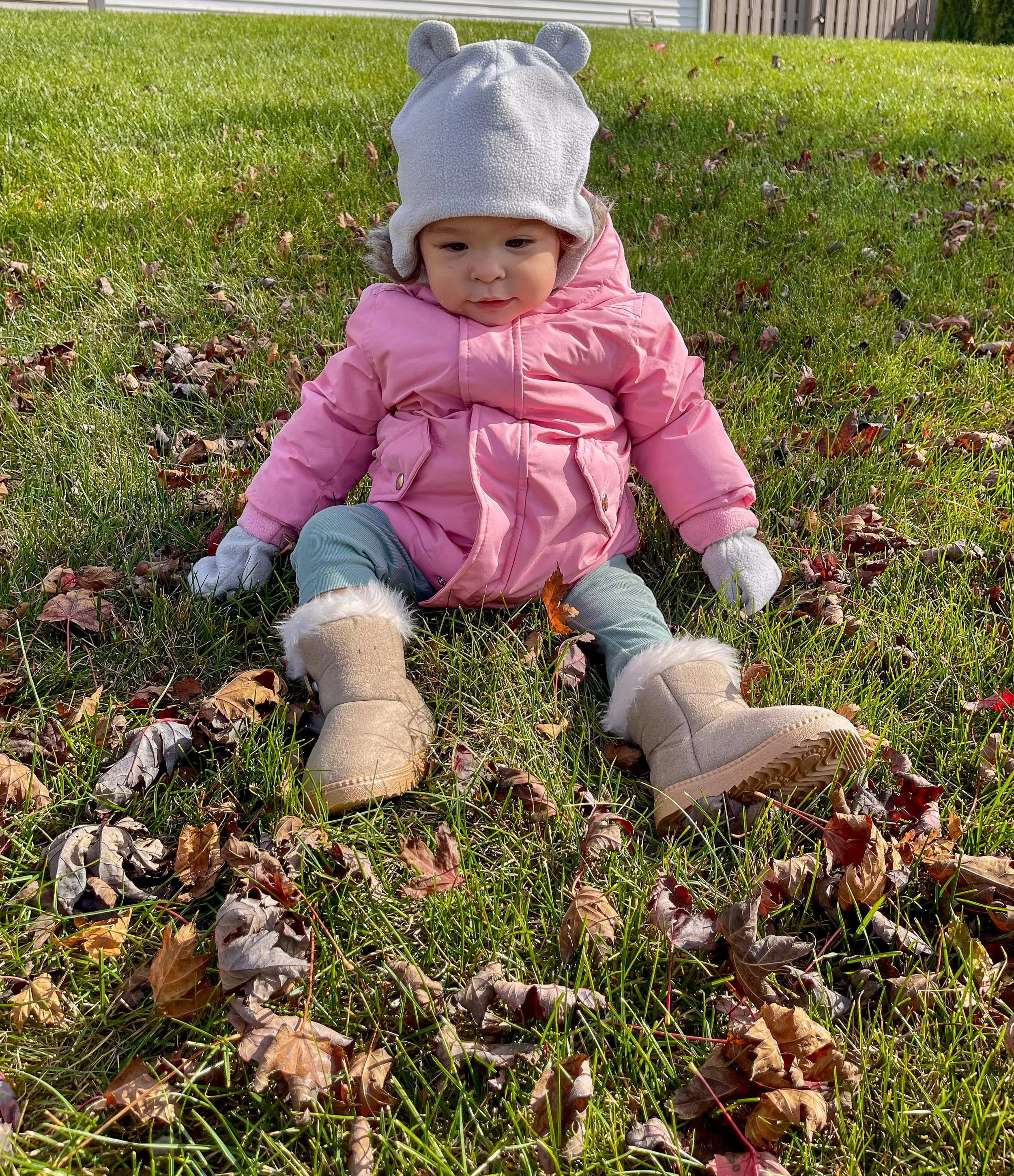 A young child sitting on grass with fallen leaves, wearing a pink coat, tan boots with fur lining, gray pants, white mittens, and a white hat with bear ears.