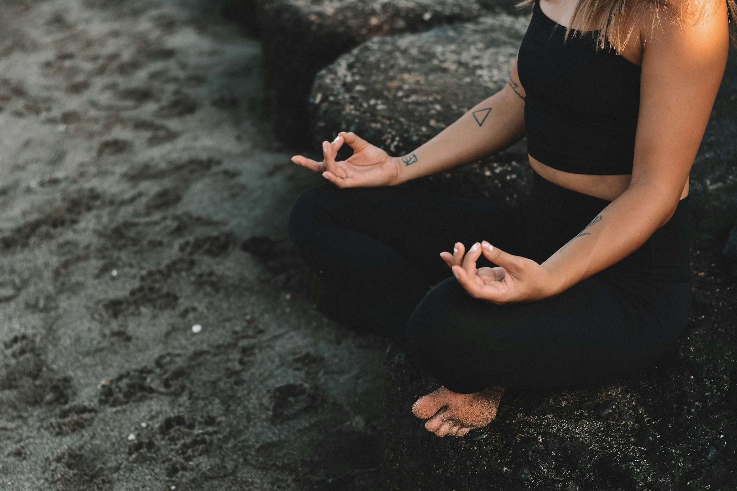 A person practicing yoga or meditation on a dark, sandy surface near rocks, with tattoos visible on their arm, wearing a black sleeveless top.