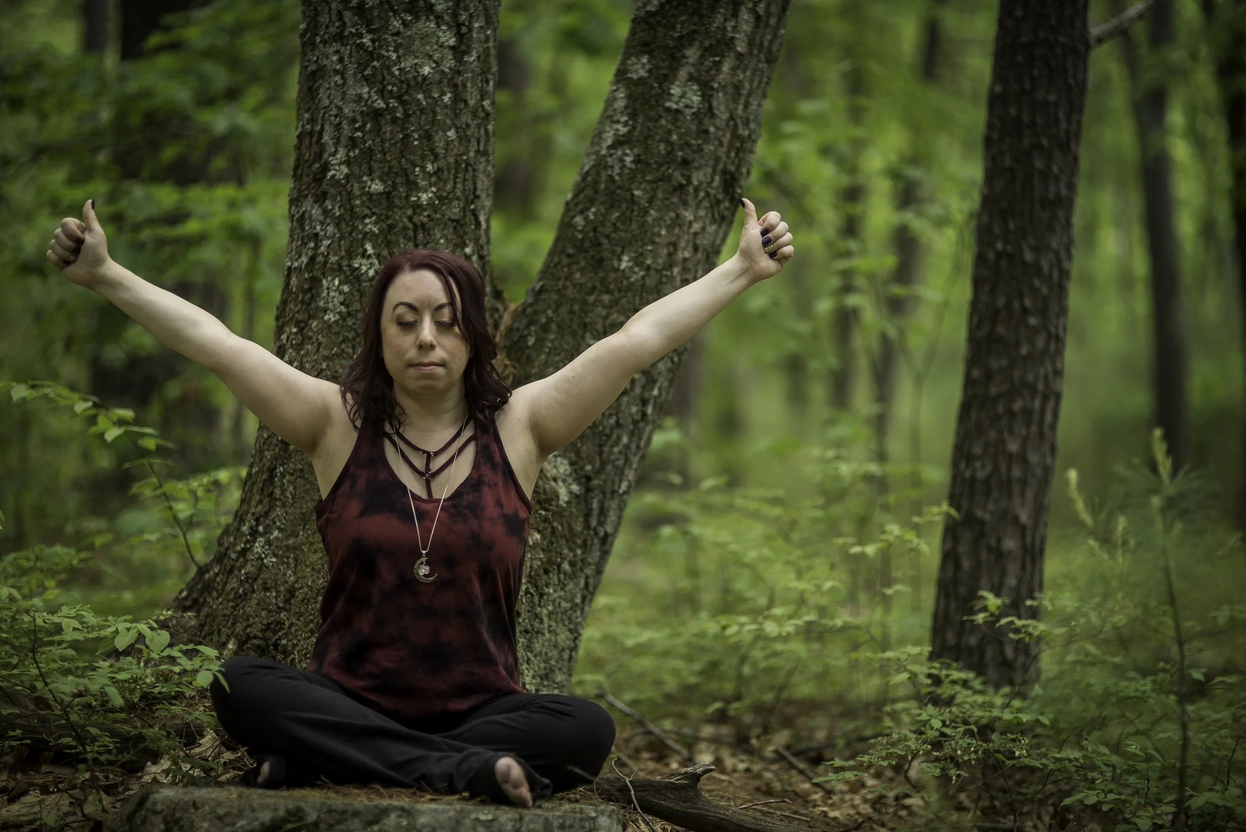 A woman with blonde long hair tied up meditation with beige pants red socks and black tank top with left hand on heart and right hand on stomach with green plant behind her