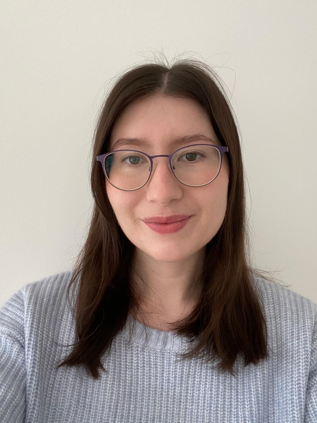 A young woman with shoulder-length brown hair, wearing round glasses with purple frames, smiling, and dressed in a light-colored, ribbed shirt against a plain white background.
