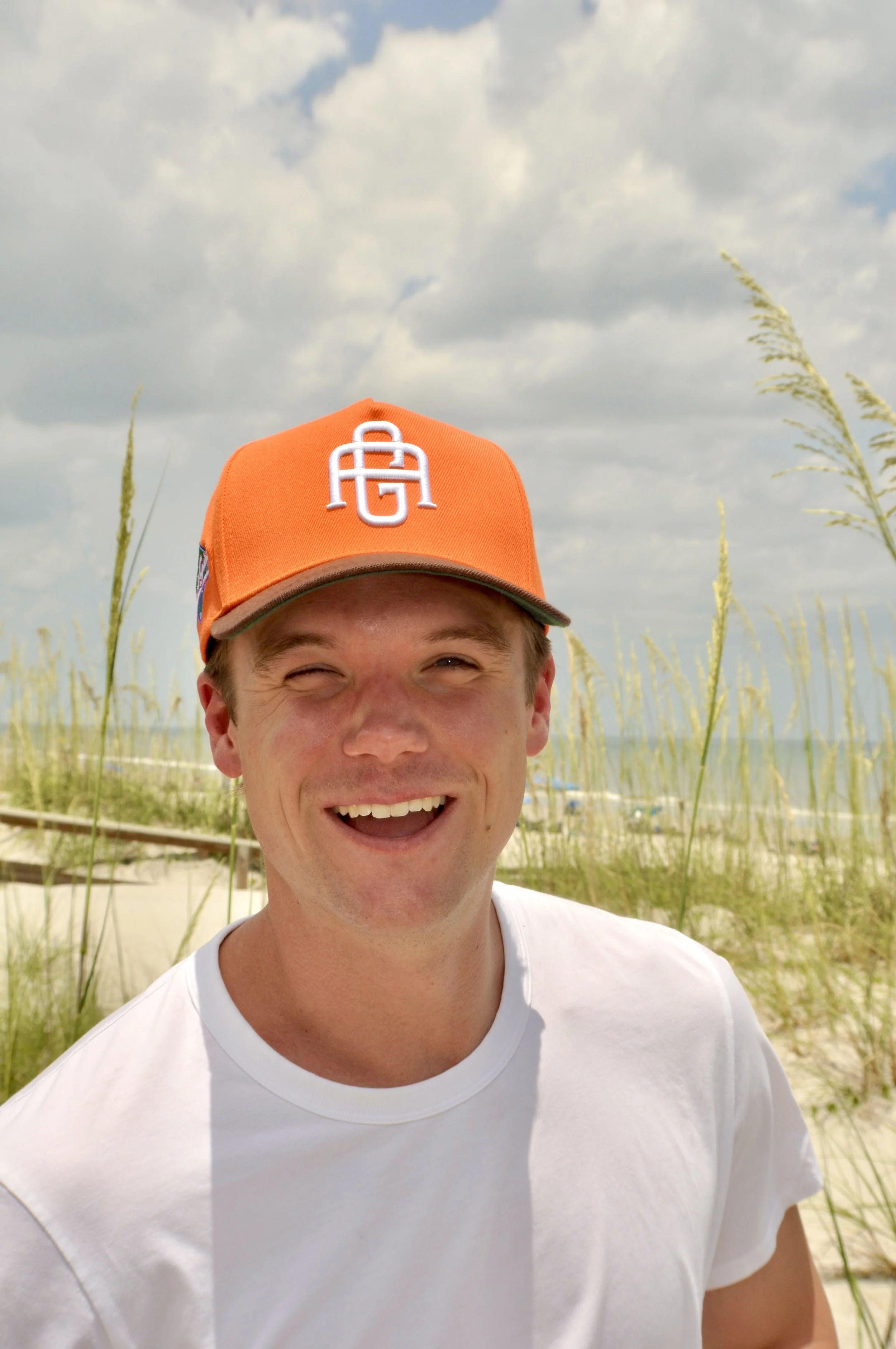 A smiling young man wearing an orange cap and a white t-shirt, standing outdoors near a beach with sand, tall grasses, and a cloudy sky in the background.