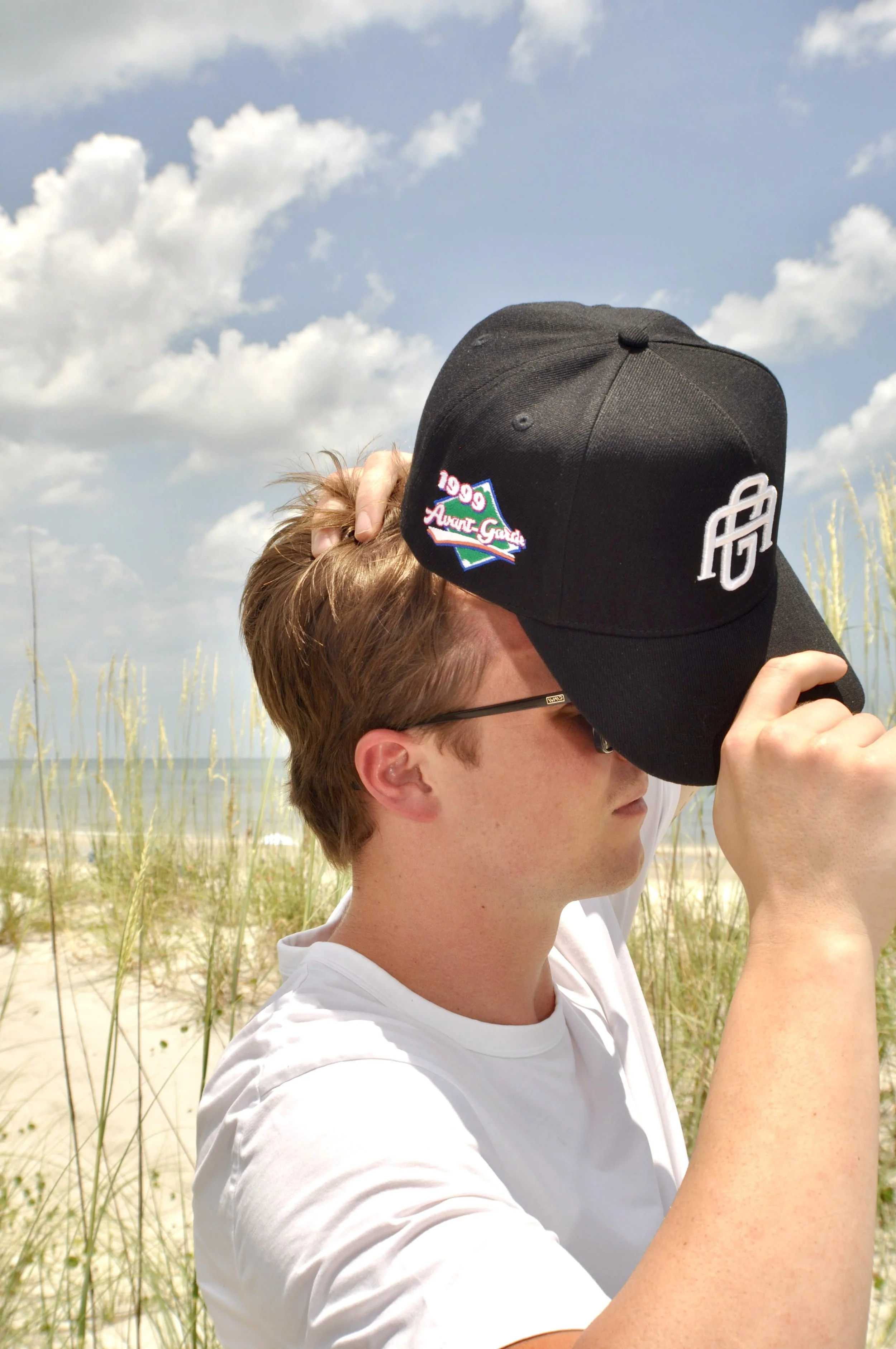 A young man with glasses in a white shirt at the beach, holding a black cap with a logo and a patch that says '1999 Avant Garde,' while adjusting the cap on his head. Blue sky with clouds and beach grass in the background.