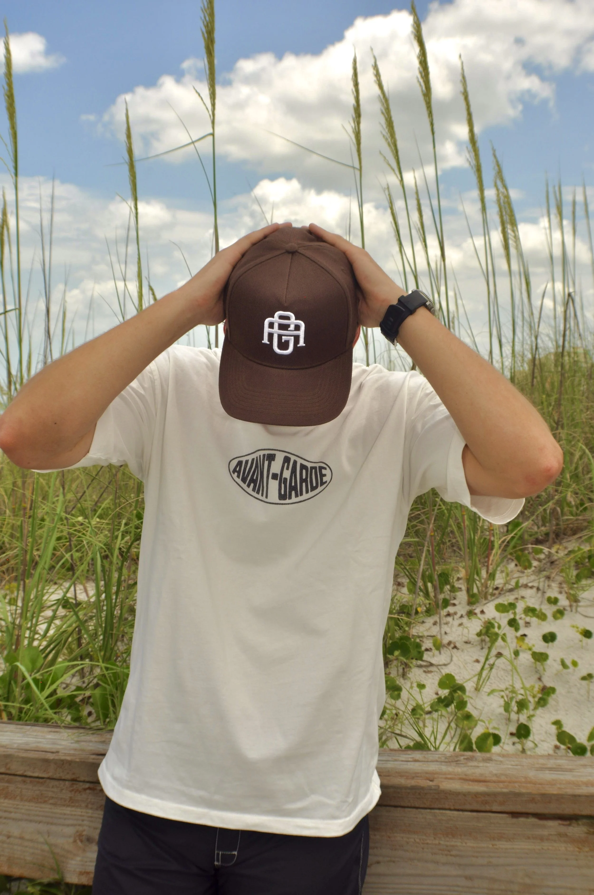 Person wearing a white t-shirt with 'AVANT-GARDE' written on it, holding a brown cap with a logo over their face, standing in front of a grassy beach with sand, green plants, and a blue sky with scattered clouds.