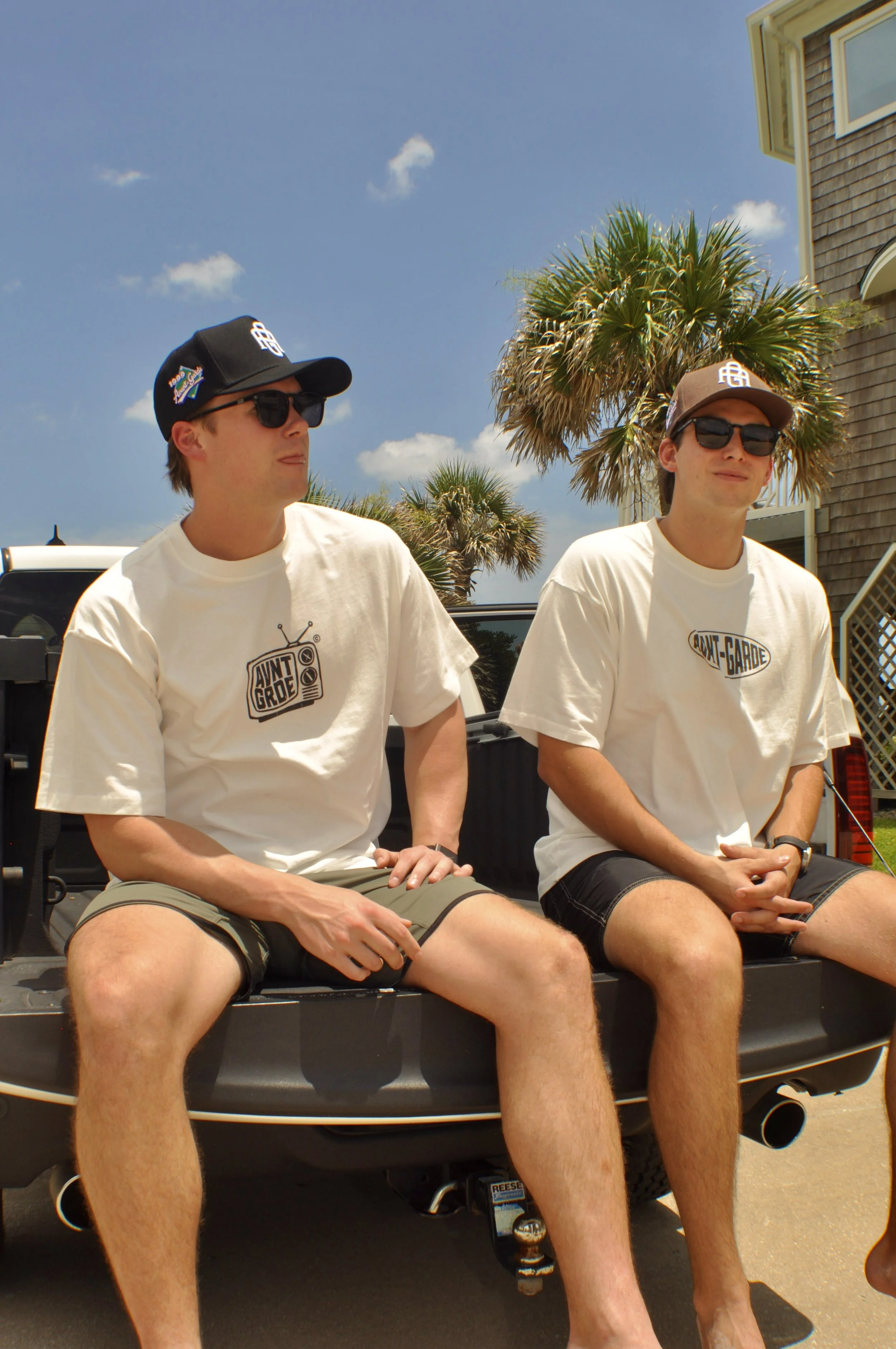 Two young men sit on the back of a black pickup truck parked outdoors under palm trees on a sunny day, wearing white t-shirts and sunglasses.