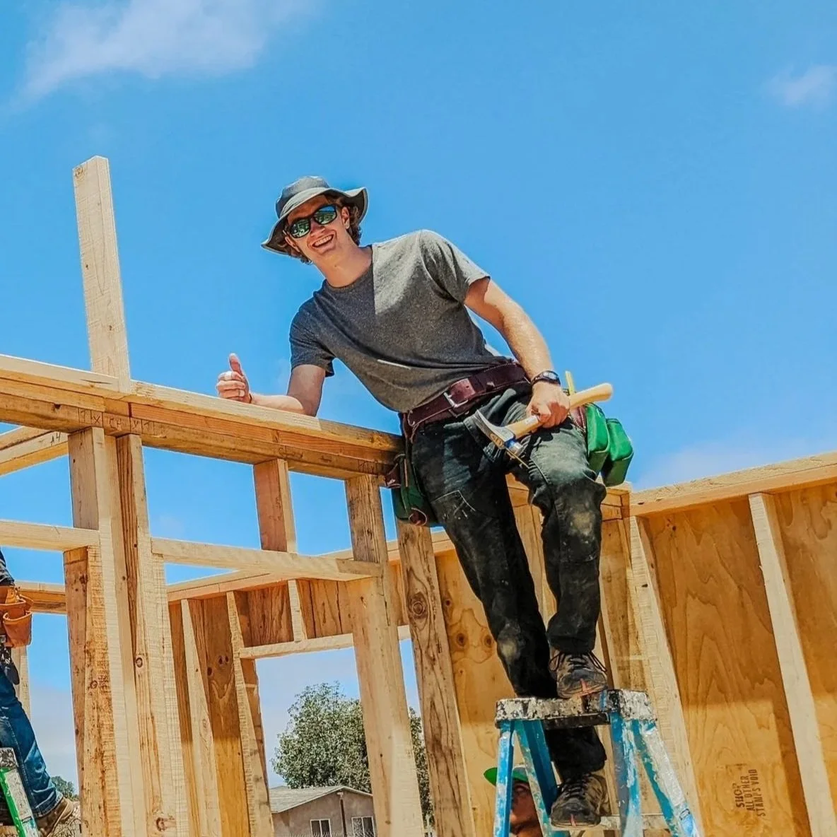 A construction worker with sunglasses and a hat smiling and giving a thumbs-up while standing on a ladder on a wooden framing project under a clear blue sky.