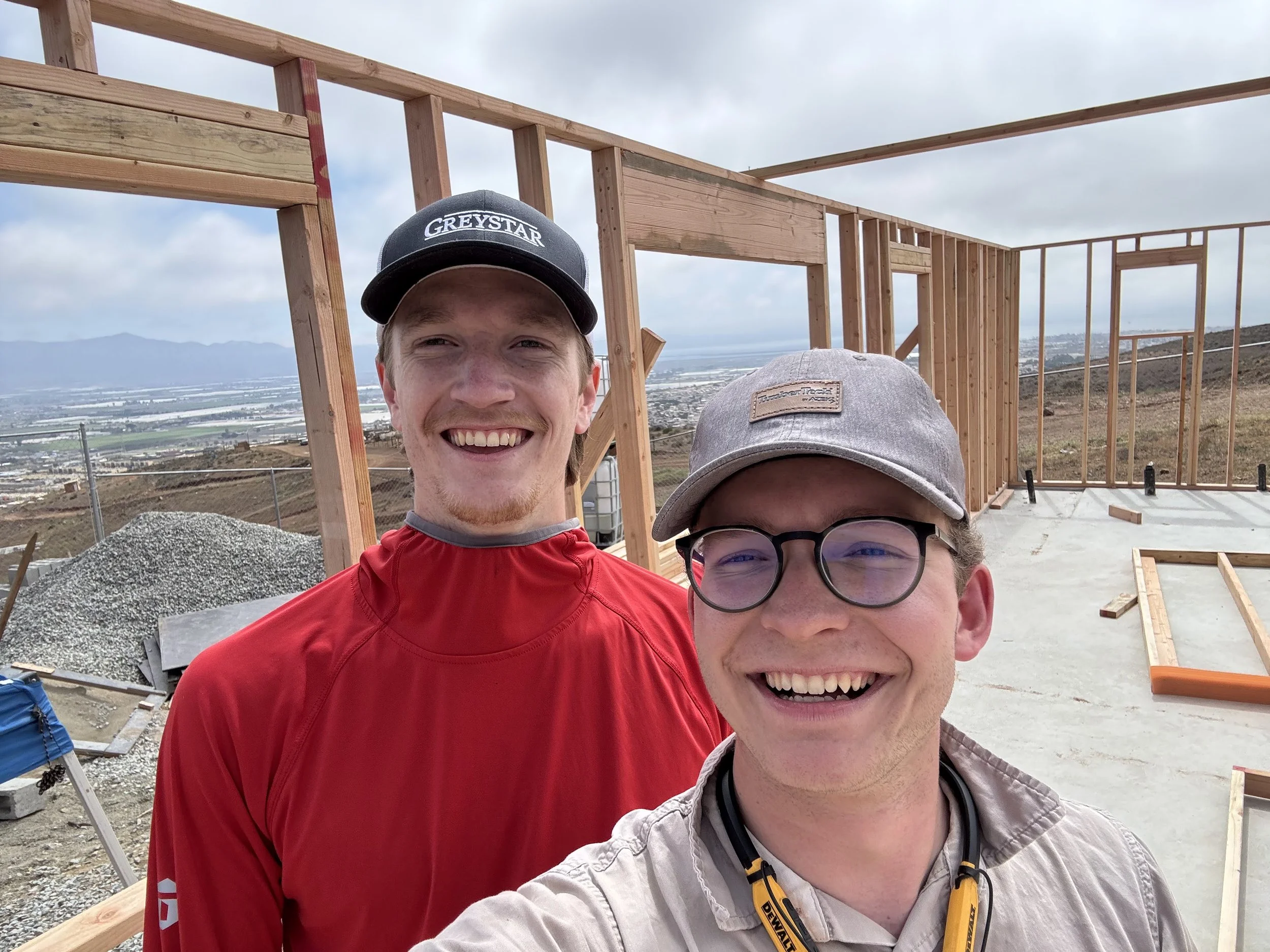 Two smiling men taking a selfie on a construction site with a wooden framework structure behind them, overlooking a city and coastal landscape under a cloudy sky.