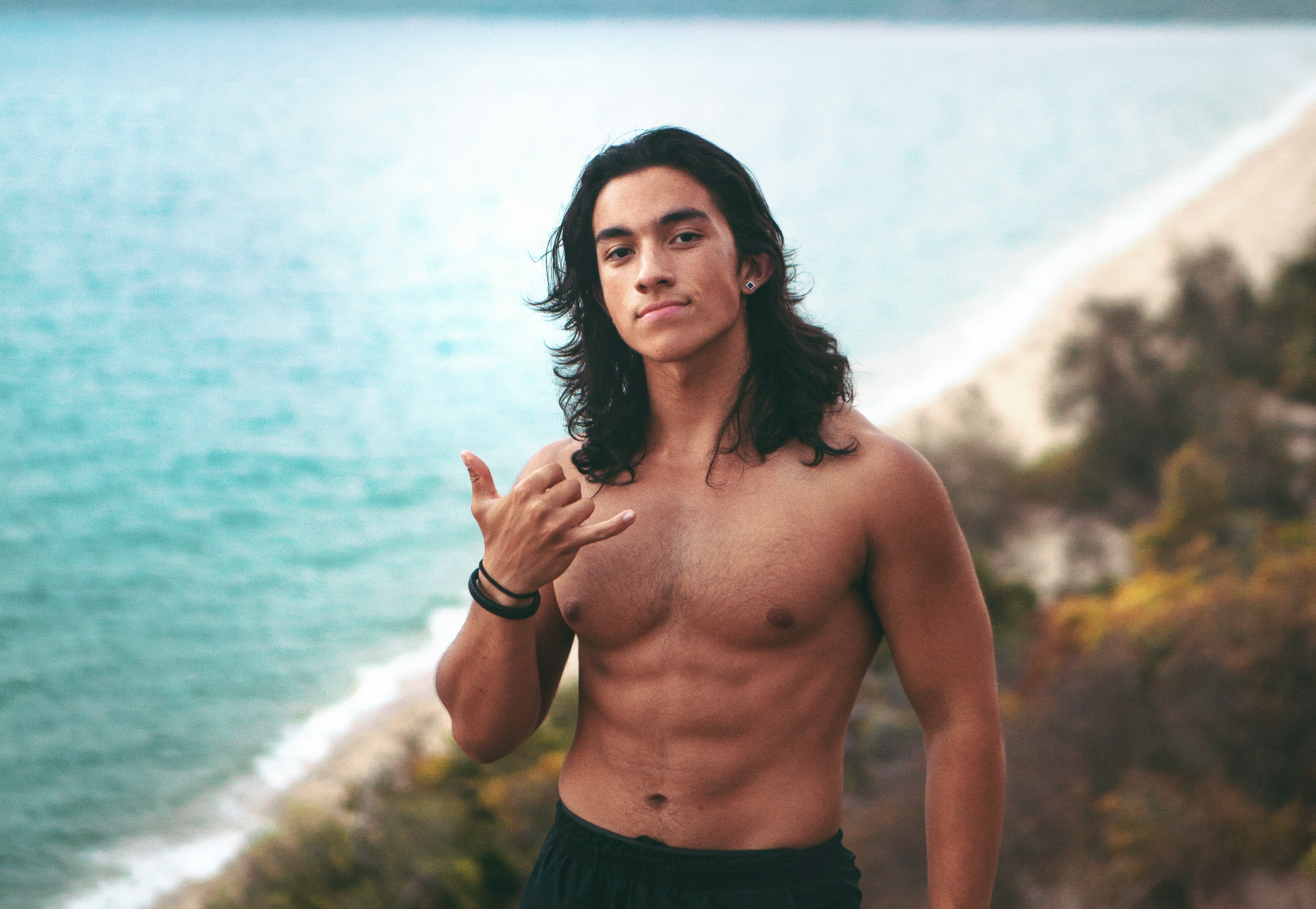 Man with long dark hair shows shaka hand signal on an overlook above a beach
