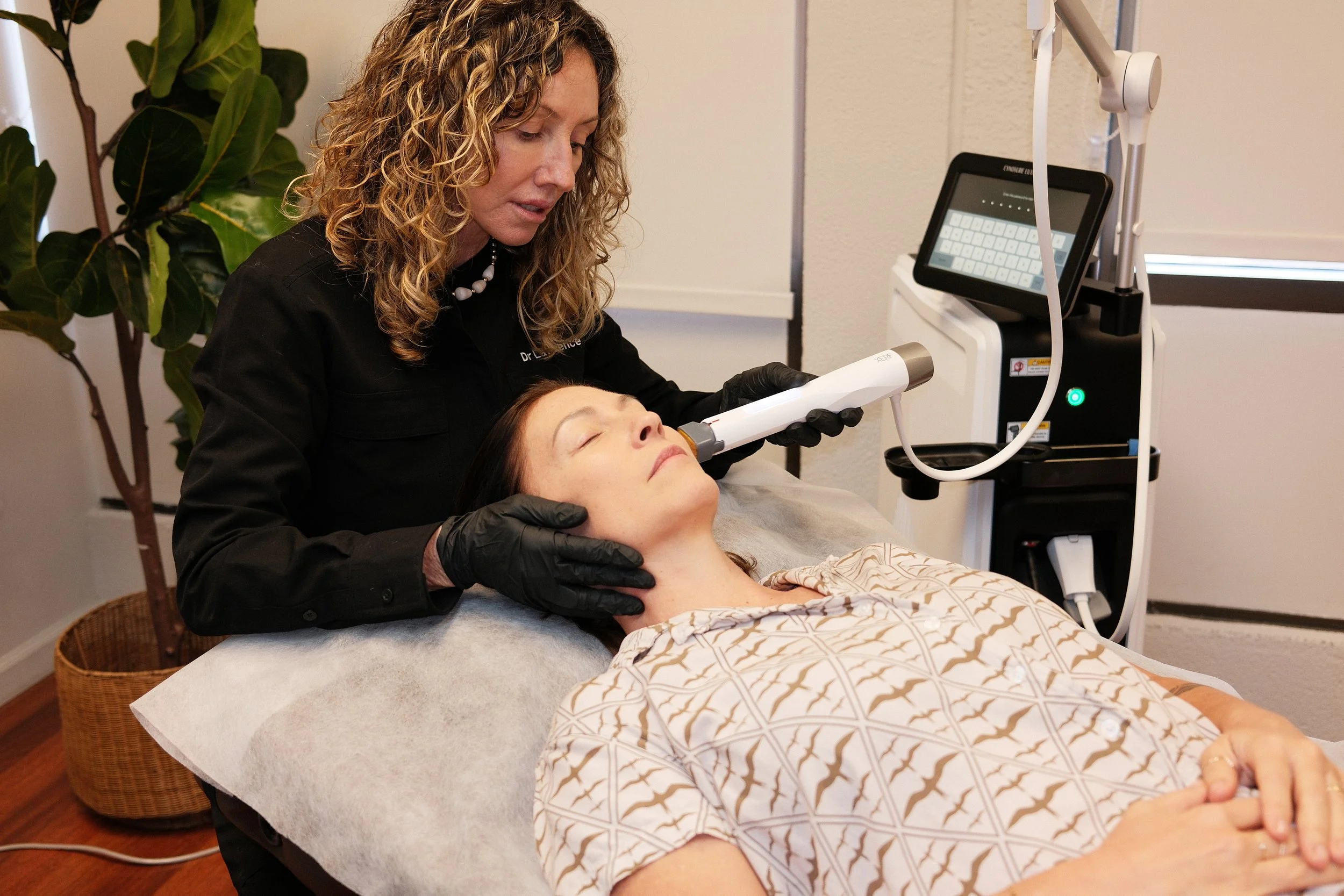 A woman lying on a medical bed is receiving a treatment with a handheld device from a healthcare professional in a clinical setting, with medical equipment nearby and a plant in the background.