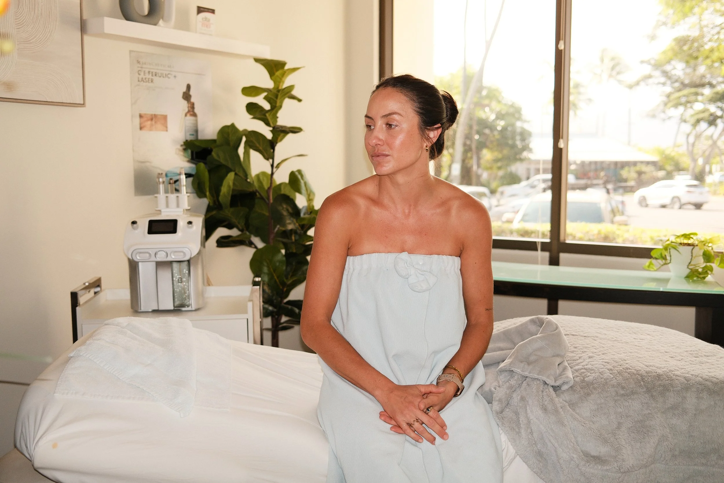 A woman sitting on a medical examination bed, wearing a white towel dress, inside a clinic or spa room with large window and green plants.