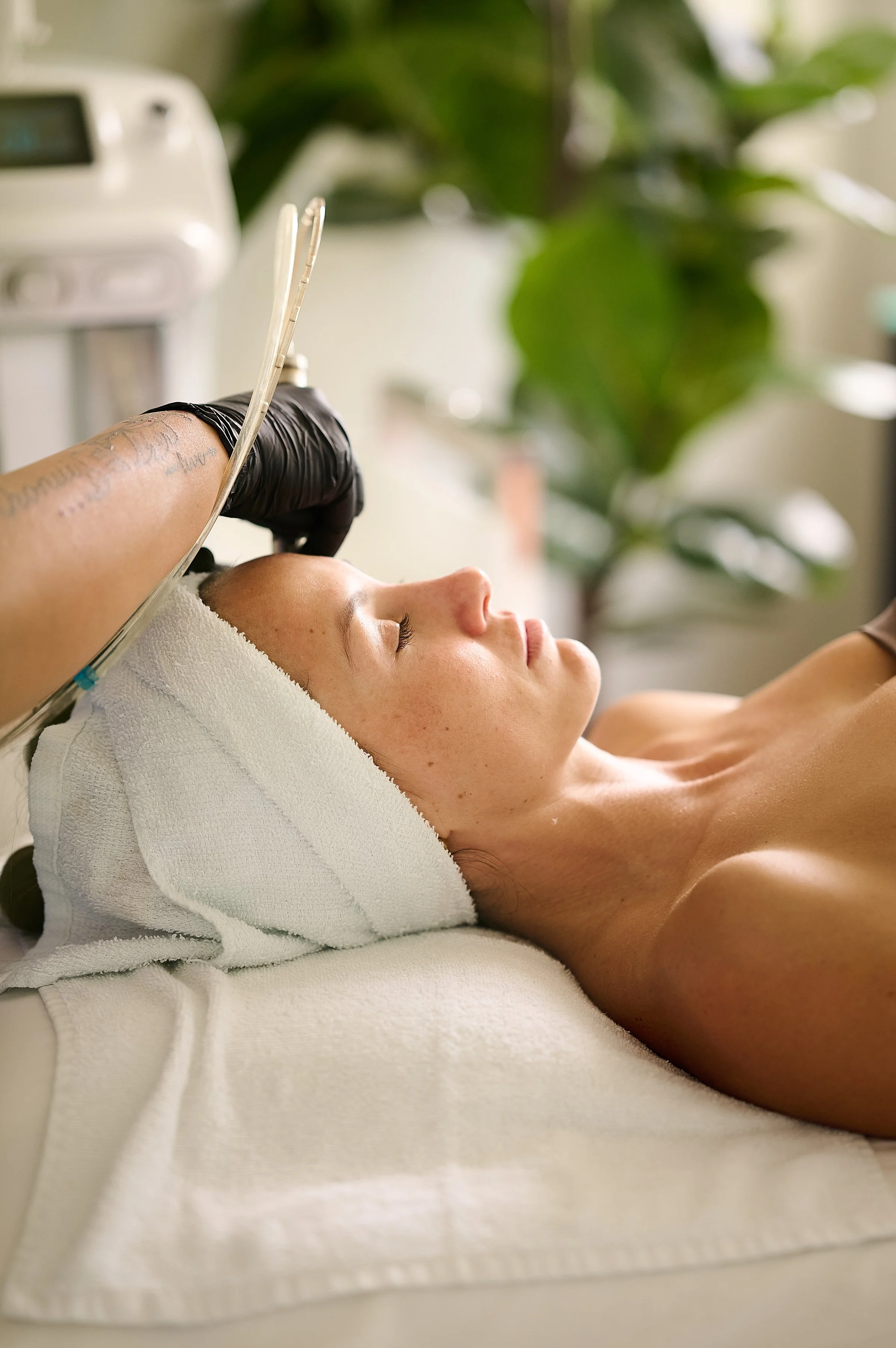 A woman with a towel wrapped around her head is lying down with her eyes closed, receiving a facial treatment from a skincare professional wearing black gloves. The setting is the Enhance Hawaiʻi medspa with green plants in the background.