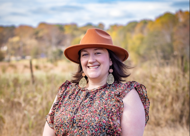 Rosamarie Gennuso smiling outdoors wearing a wide-brimmed brown hat, floral blouse, and dangling earrings, with trees and a cloudy sky in the background.