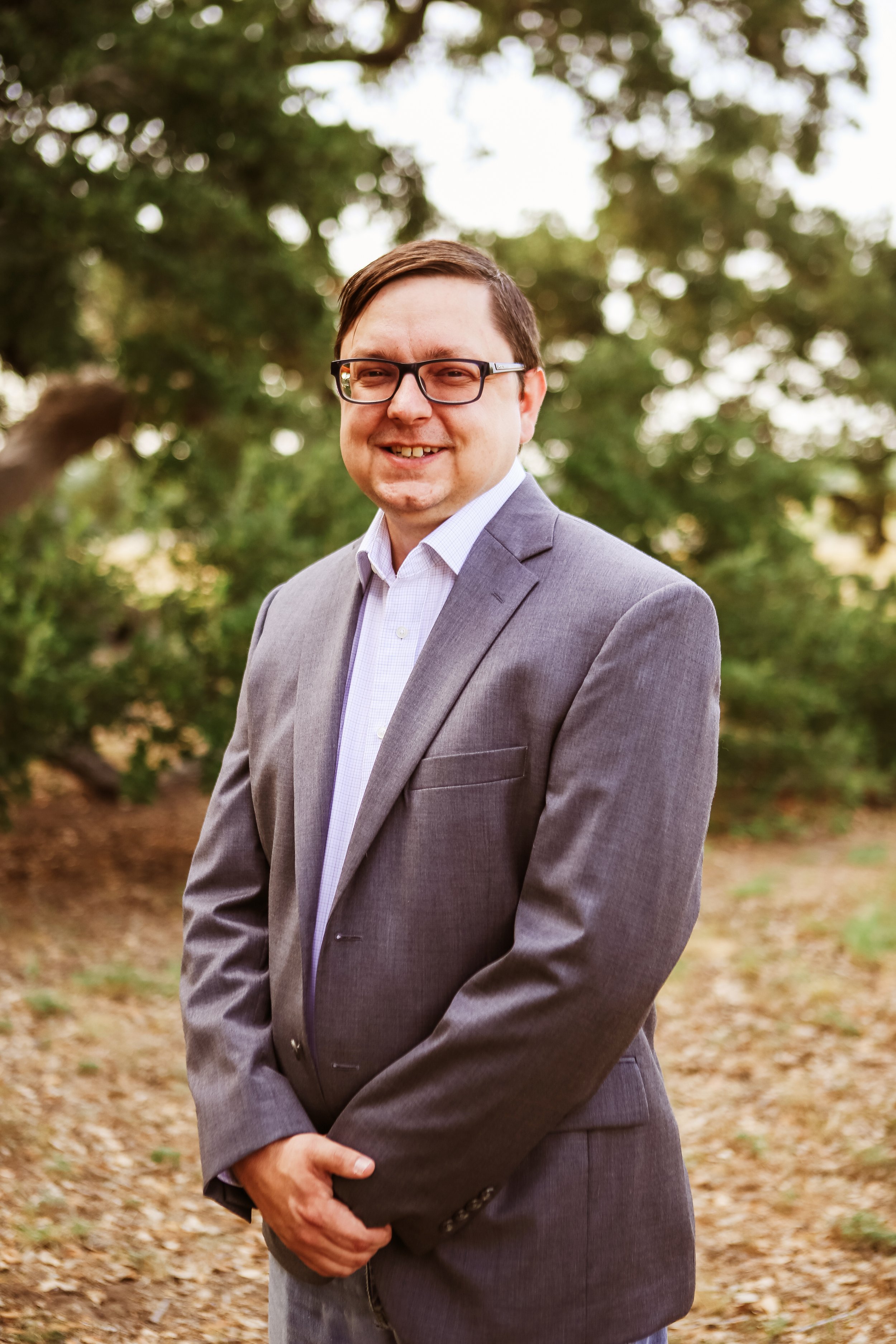 Gerry Hogsed in a grey suit and white shirt standing outdoors among trees, smiling.