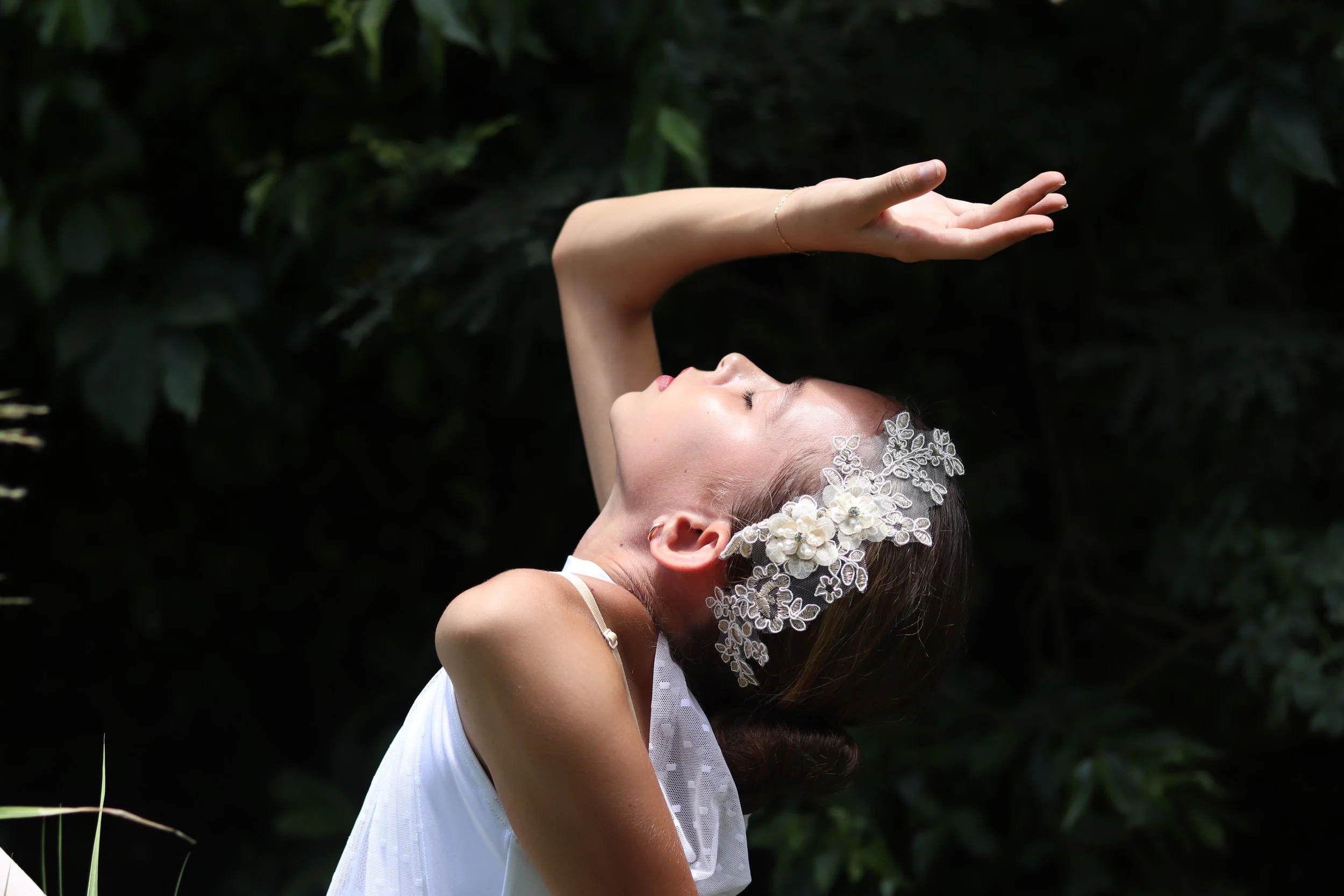 A young woman in a white dress with a floral headpiece posed outdoors with her eyes closed, head tilted back, and one arm raised above her head against a dark green leafy background.