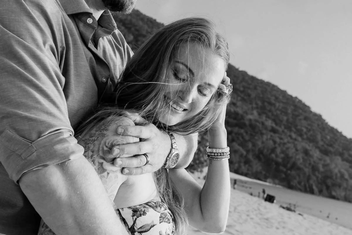 A couple on a beach, with the man embracing the woman from behind, both smiling with eyes closed, and a hillside in the background.