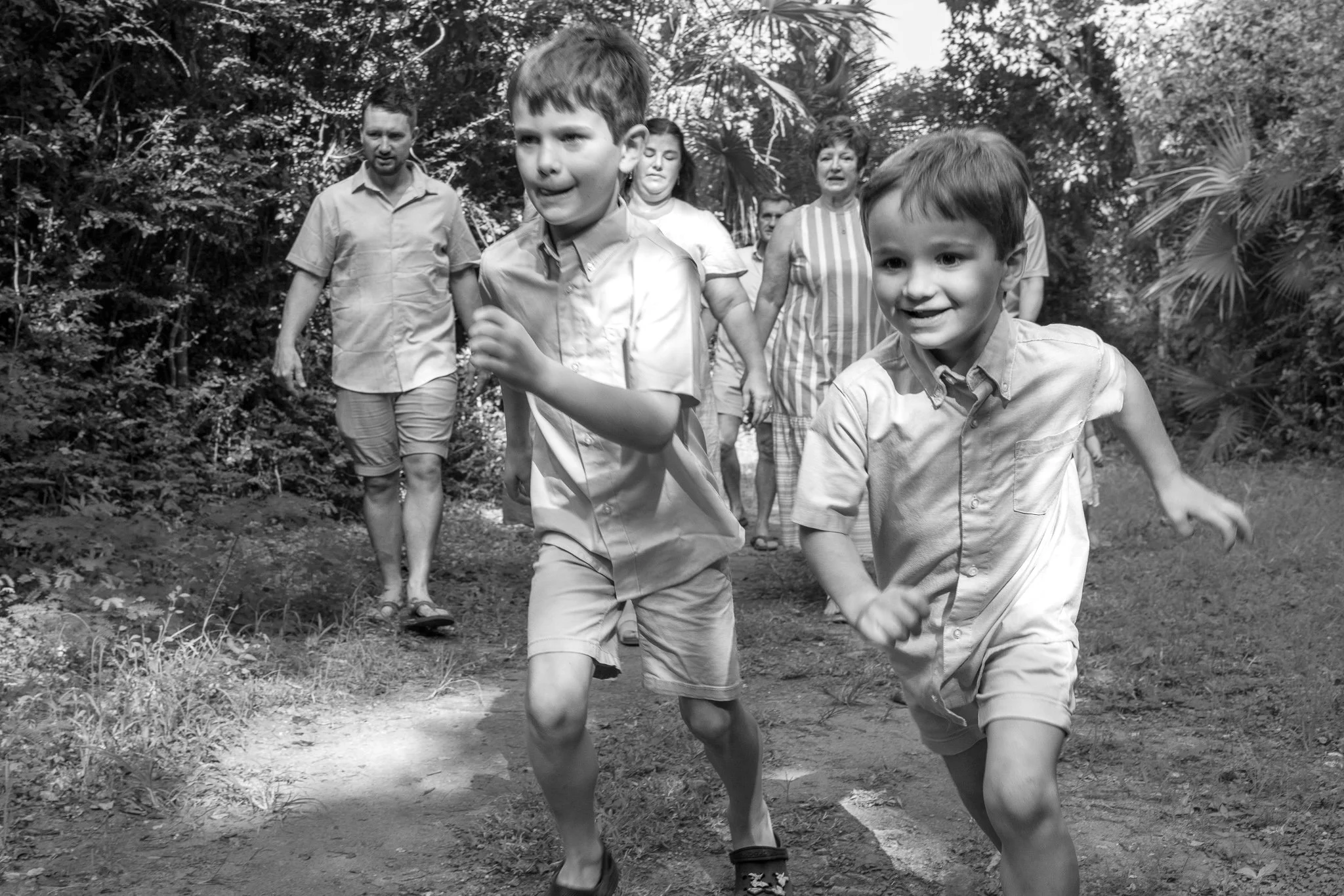 A group of children and adults running outdoors on a dirt path surrounded by trees and bushes. The image is in black and white.