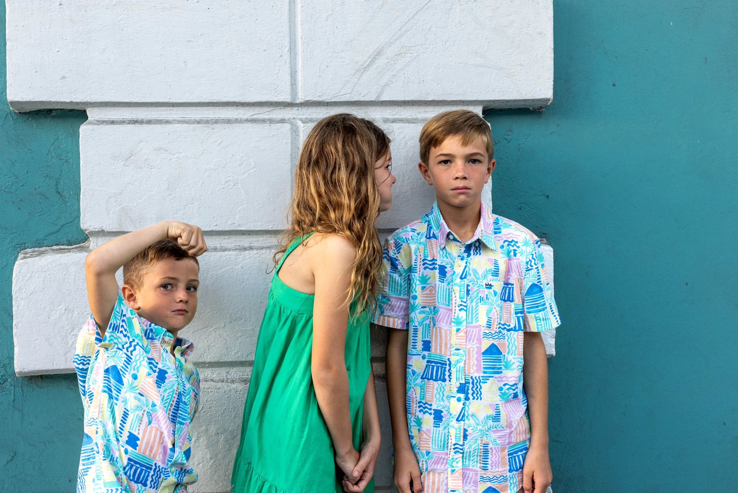 Three children standing against a wall, with the girl whispering to the boy on the right, and the younger boy on the left striking a pose.