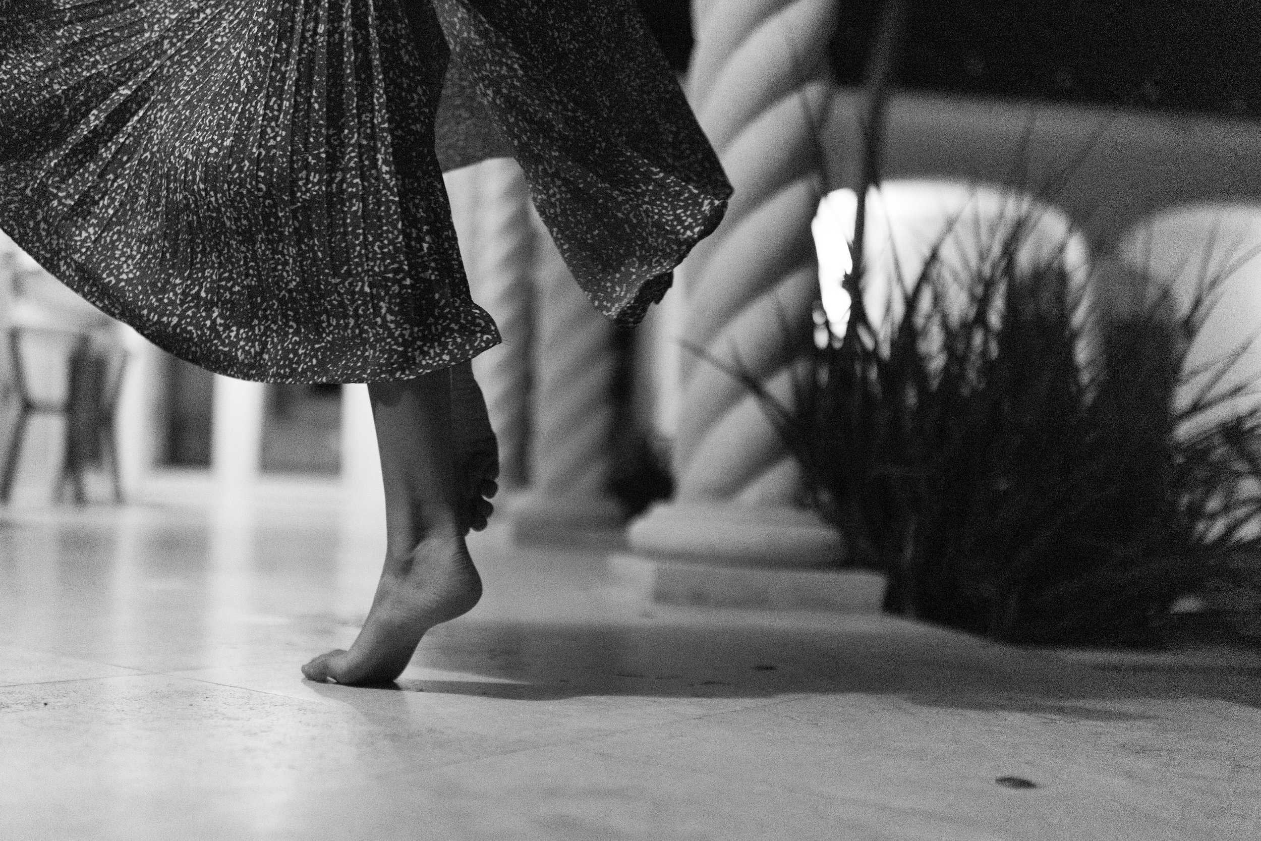 A person is standing on tiptoe on a wooden floor, with their back slightly bent. They are wearing a lightweight, textured, patterned dress with flowing sleeves. In the background, there are interior elements like a chair and a potted plant.