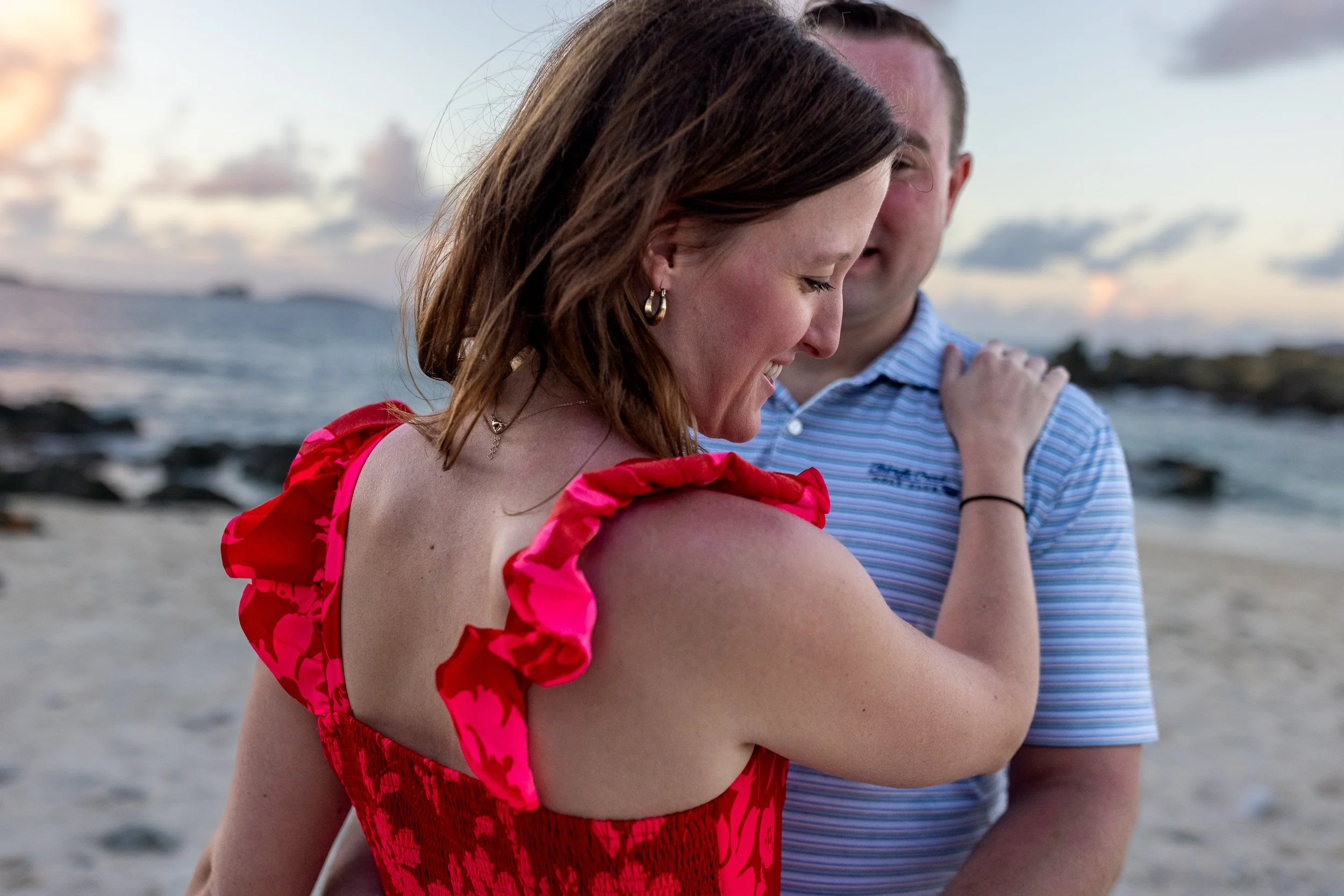 A young woman in a red dress with ruffled sleeves smiling and looking down, with a man standing behind her on a beach at sunset, with the ocean and distant islands in the background.