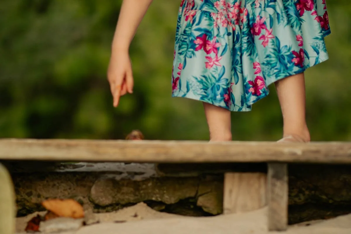 Close-up of a child standing on a wooden bridge with a floral dress, with the background blurred greenery.